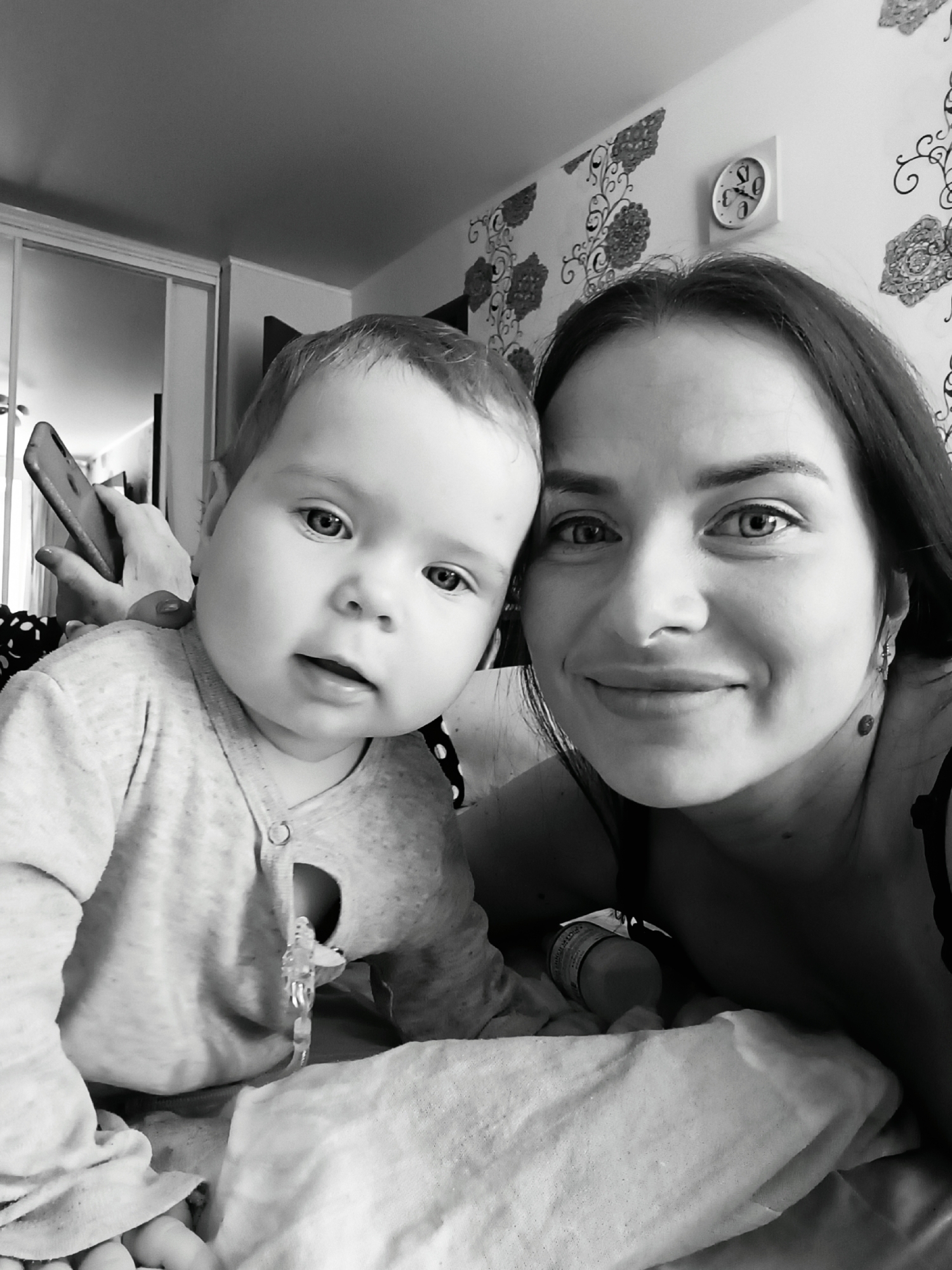 The image shows a woman and a baby, likely indoors. The woman is on the right side of the frame, looking at the camera and smiling. The baby is on the left, also looking towards the camera. The background consists of a wall with patterned wallpaper, a clock showing approximately 7:05, and part of a sliding door wardrobe. The baby wears a light-colored, buttoned outfit with a clear object attached. There is a bottle of pills on the bed near the woman. A hand holding a phone is visible behind the baby. The location is Chaikovsky, Russia.