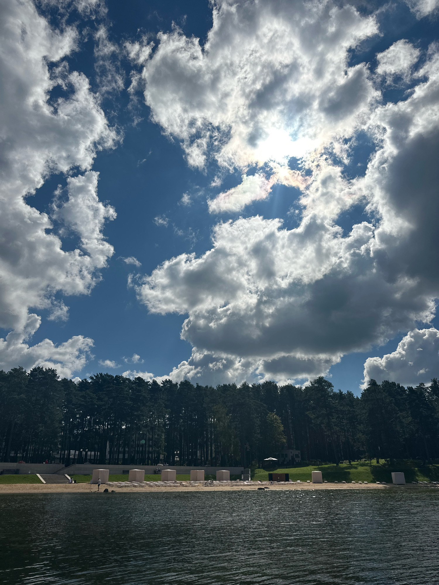 The image depicts a scenic view of a lake in the Мядзельскі сельскі Савет, Belarus. The foreground is dominated by the dark, rippling surface of the lake. Beyond the lake, a sandy beach stretches across the midground, lined with numerous empty lounge chairs and a few scattered beach structures. A dense forest of tall, dark green trees forms a solid barrier along the edge of the beach. The sky above is a vibrant blue, filled with a dramatic display of white and gray cumulus clouds. The sun is visible, peeking through a gap in the clouds, casting a bright light and creating lens flares. The overall impression is one of a serene, natural landscape, likely a resort or recreational area, under a dynamic sky.