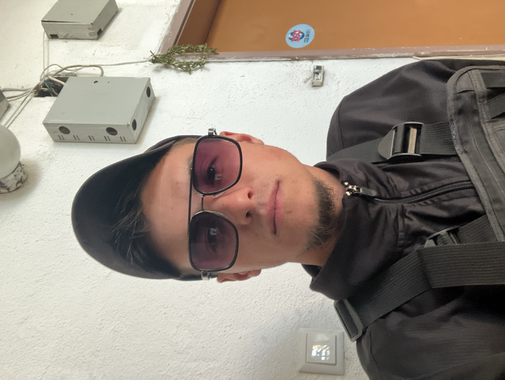 A man is seen in a close-up selfie taken at a low angle. He is wearing sunglasses and a black cap, and a black backpack is slung over his shoulder. The background consists of a white wall with electrical boxes and conduit, as well as a doorway with a sticker on the doorframe. The location is Plovdiv, Bulgaria.