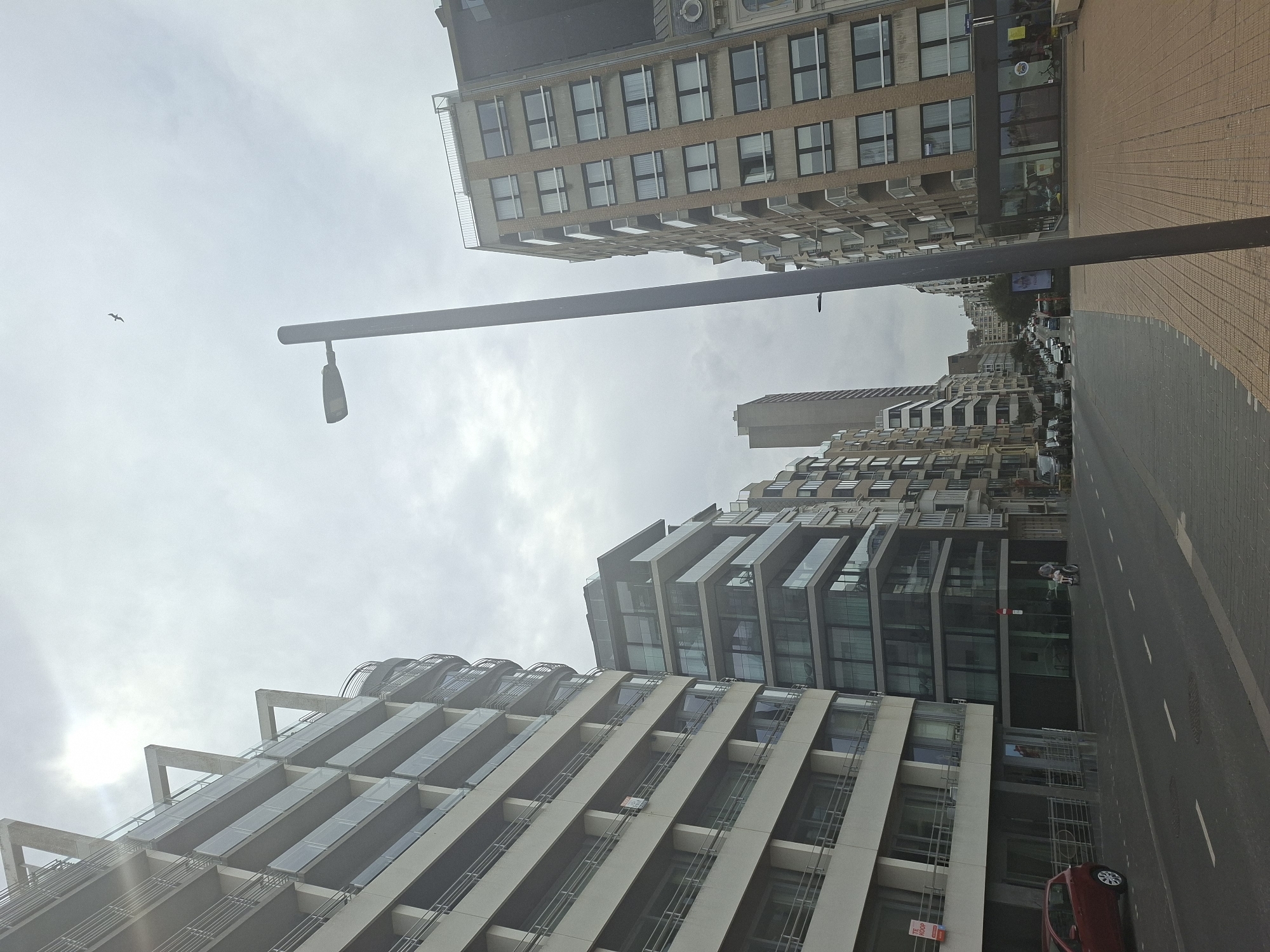 This image depicts a street scene in Ostend, Belgium, likely along the coast, given the architectural style of the buildings. The perspective is from street level, looking down a street lined with multi-story apartment buildings and businesses. The sky is overcast with some bright patches, indicating it's daytime, and a bird is visible flying in the distance.
On the left, a large, light-colored apartment building with multiple balconies dominates the foreground. A red car is parked on the street in front of this building. Further down the street, several other modern apartment buildings with glass balconies and facades are visible, along with older, more traditional buildings.
The street itself is paved, with a pedestrian walkway made of brown and grey tiles. Traffic is present, with several cars and a person on a scooter visible on the road. A tall, dark lamppost stands prominently in the mid-ground, obscuring some of the view of the buildings behind it. The overall impression is of a well-maintained urban environment.