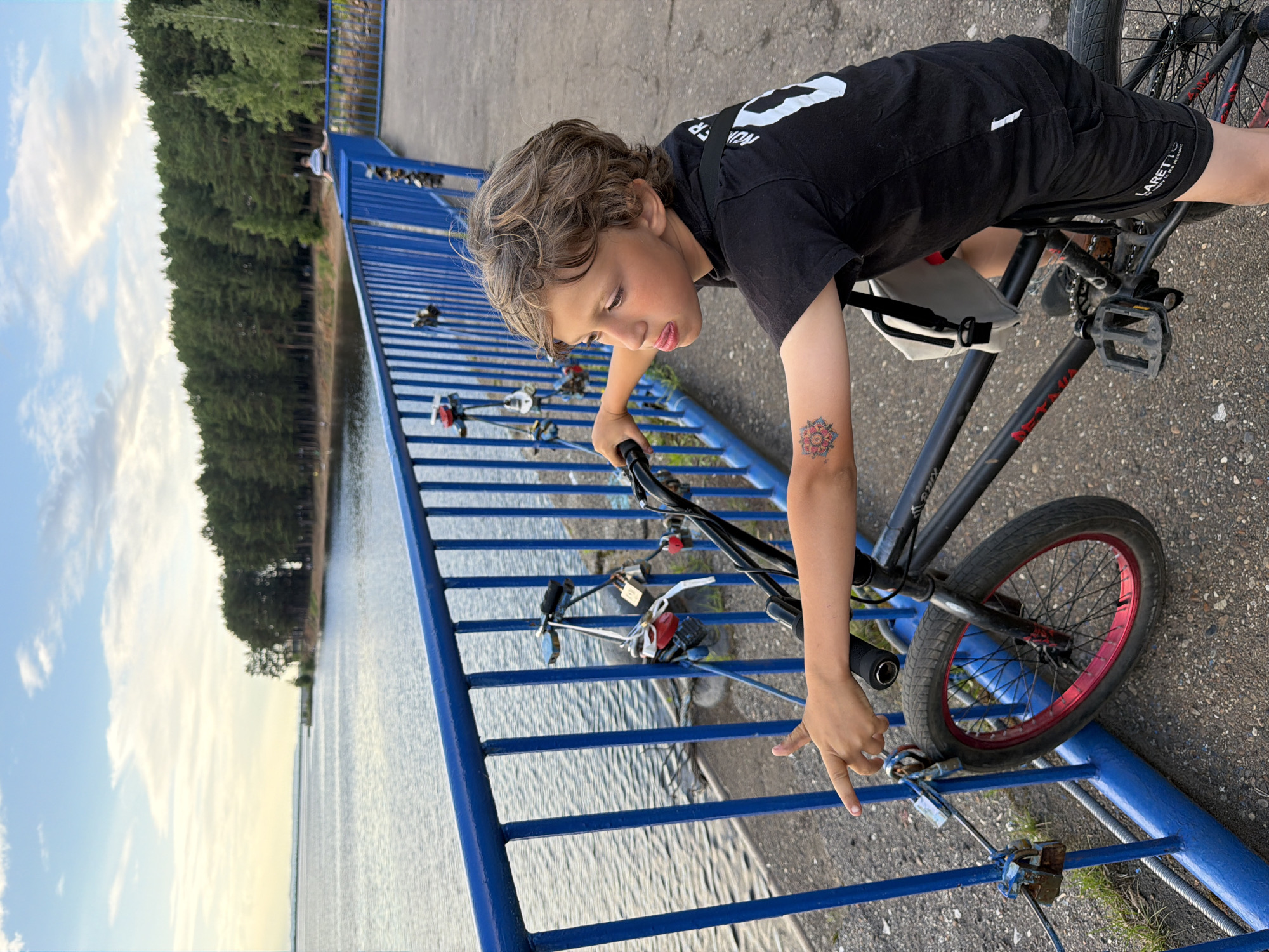 A young boy is captured in a close-up shot as he leans over the handlebars of a BMX bike. He is wearing a black t-shirt with white lettering and black shorts. A small, light-colored bag is attached to the bike's frame. The boy has curly brown hair and a tattoo of a red and pink mandala on his left arm.
The bike is parked on a paved surface next to a blue metal railing. The railing is adorned with numerous padlocks, suggesting it might be a bridge or a spot where people express their love or make wishes. Beyond the railing is a body of water, likely a lake or river, with trees lining the far bank. The sky is partly cloudy with a mix of blue and white. The lighting suggests it is daytime, possibly late afternoon or early evening. The overall scene appears to be outdoors in a recreational area. The boy's gaze is directed downwards, and his expression could be interpreted as focused or thoughtful. The text visible on the boy's shorts reads "LARETTO".