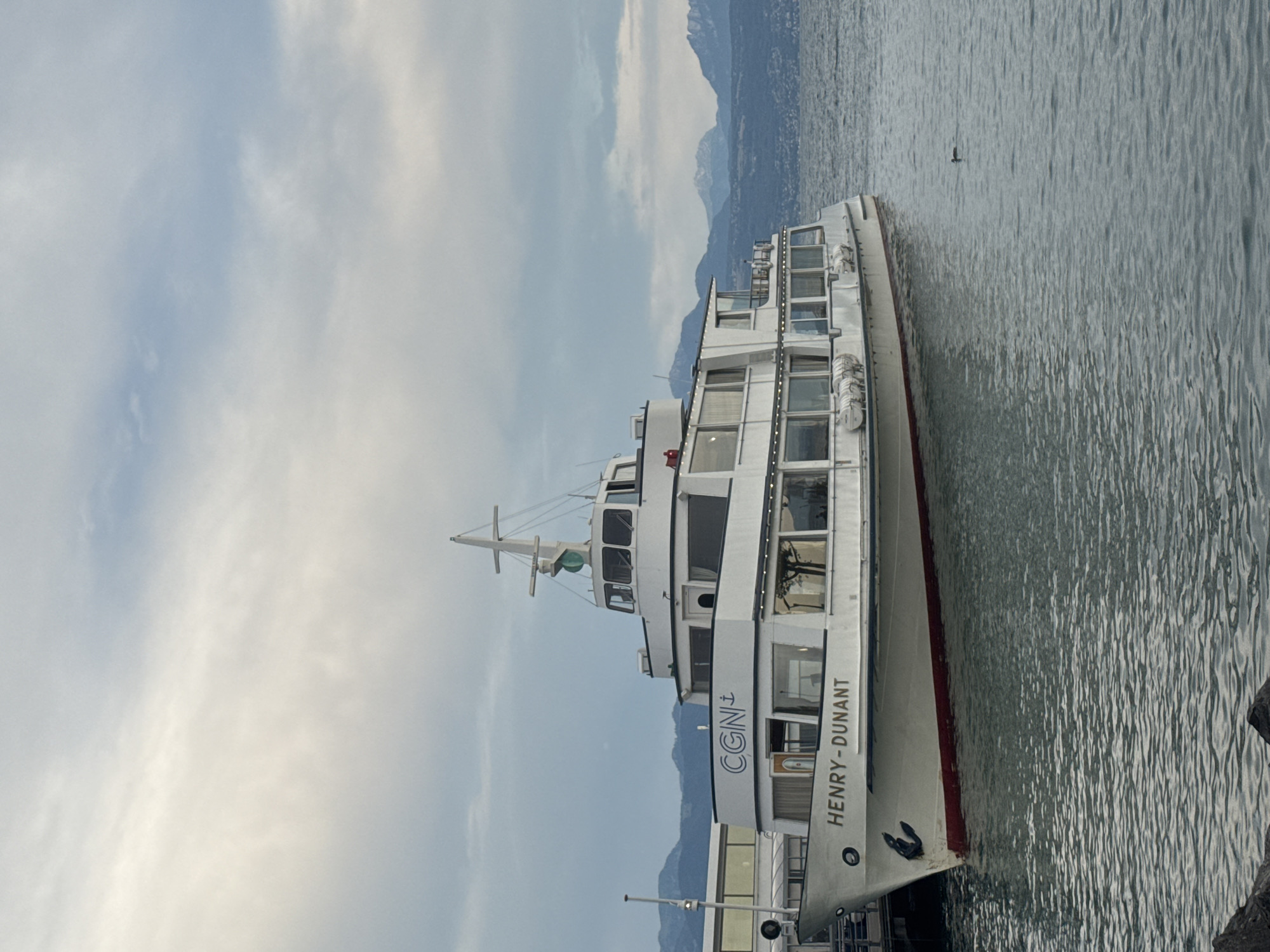 This is a full shot photograph of a large white passenger boat named "HENRY-DUNANT" docked on the left side of the frame. The boat has multiple decks and numerous windows. The name "CGN" is visible on the side of the boat above "HENRY-DUNANT," indicating it belongs to the Compagnie Générale de Navigation sur le lac Léman, a Swiss company operating public transport on Lake Geneva. In the background, there is a body of water, likely Lake Geneva, with mountains visible in the distance. The sky is overcast with clouds. The boat is not in motion, and no people are visible. The scene suggests a calm day at a port in Lausanne, Switzerland, given the CGN markings.
