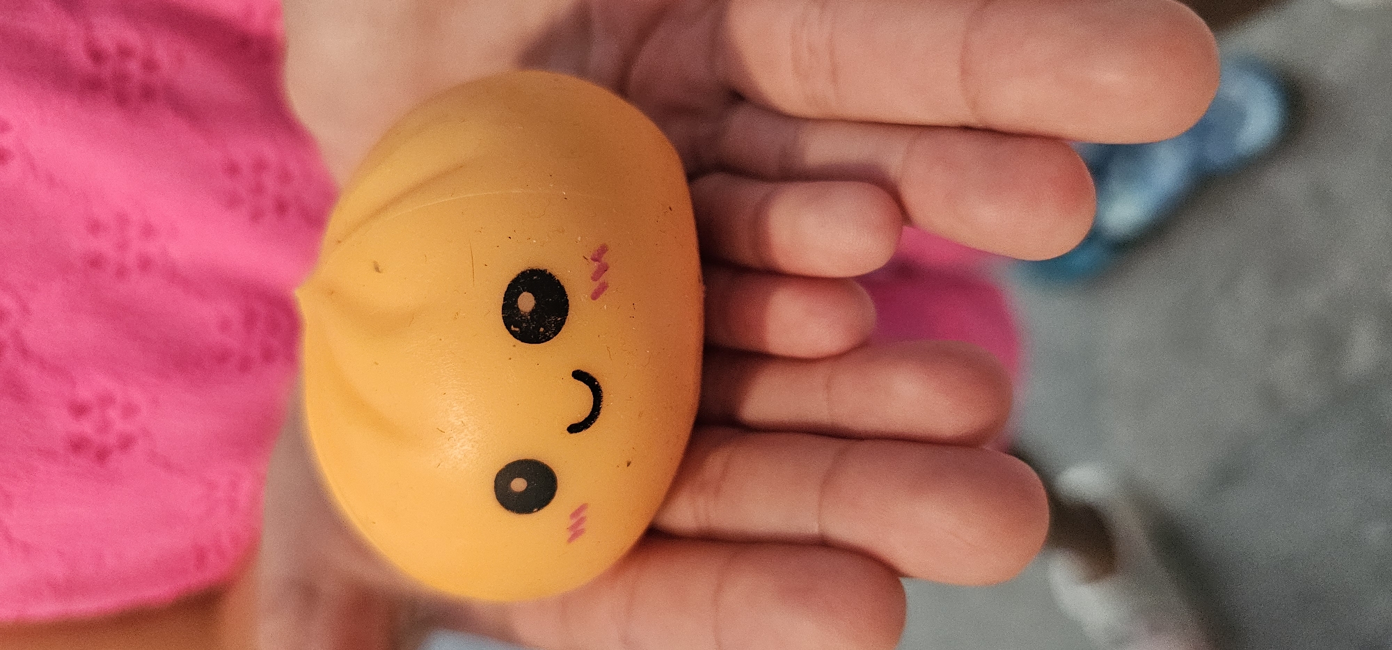 A close-up, top-down shot shows two hands holding a small, orange, bun-shaped toy with a smiling face. The toy has black eyes and a curved mouth, with pink blush marks on its cheeks. The hands are a pale skin tone, with visible fingers and fingernails. The background is out of focus and appears to be a patterned pink fabric and a grey surface. The image is sharpest on the toy and the hands. The image does not contain any identifiable landmarks or contextual information that would allow for its location to be specified within the city of Grad Supetar, Croatia.