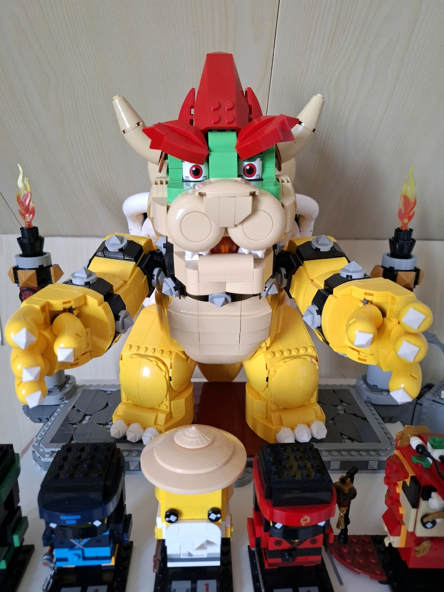 The image displays a collection of Lego figurines arranged on a shelf against a neutral, wood-grained background. The central and largest figurine is a detailed Lego model of Bowser, the antagonist from the Super Mario franchise, characterized by his yellow body, red and green head accessories, and large claws. To the left and right of Bowser are two Lego torches with flames. In the foreground, several smaller Lego minifigures are visible, including at least two figures resembling characters from Ninjago. There is no discernible indication of the city of Tyumen, Russia, nor any information pertaining to its location within that city. The scene is static and depicts a display of toys.