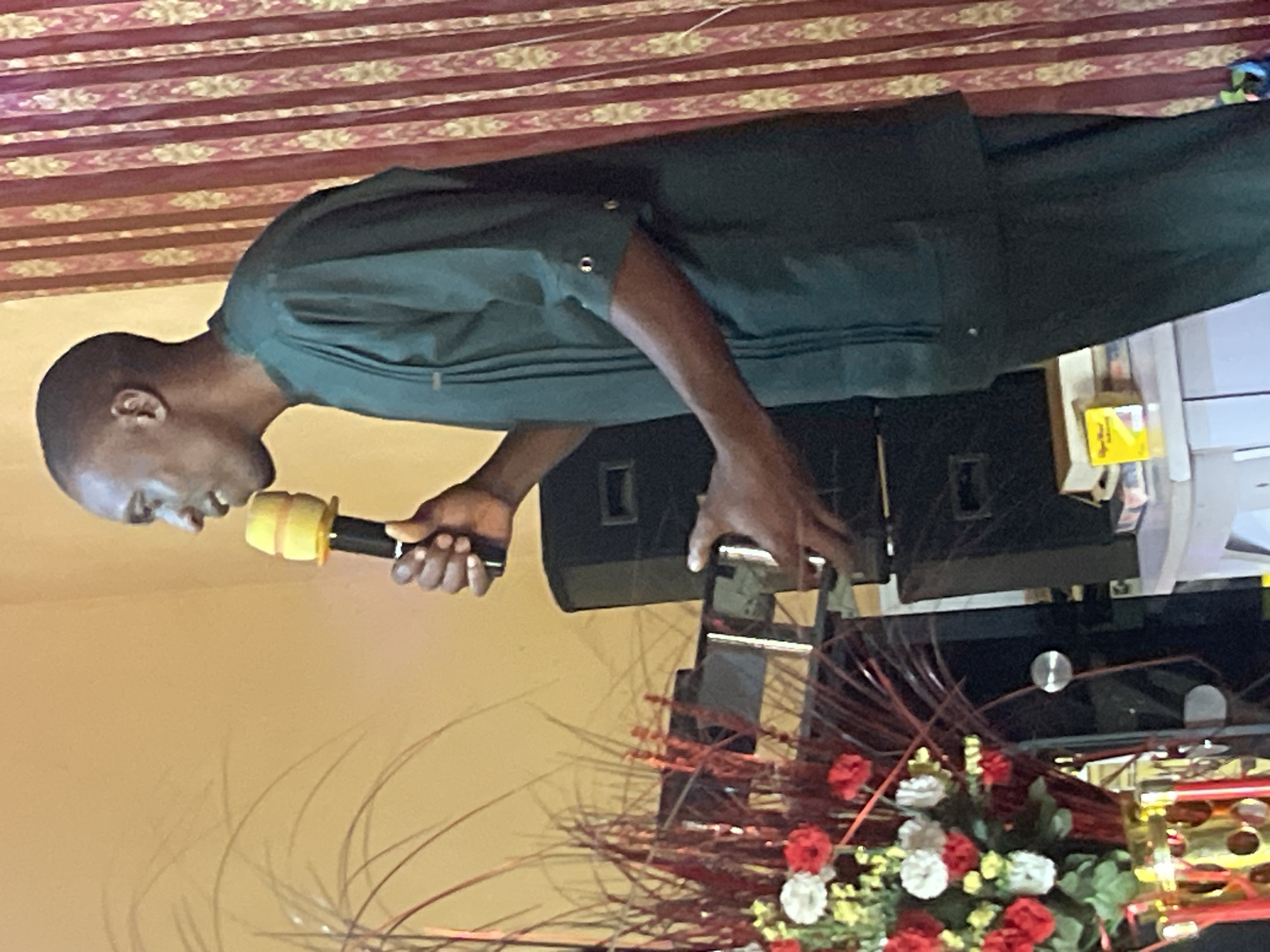 A man is speaking into a microphone with a yellow-colored windscreen. He is wearing a dark green long-sleeved shirt. Behind him are speakers, and in front of him, there is a flower arrangement with red and white flowers. The background has a red and gold pattern. The location is Unknown, Unknown.