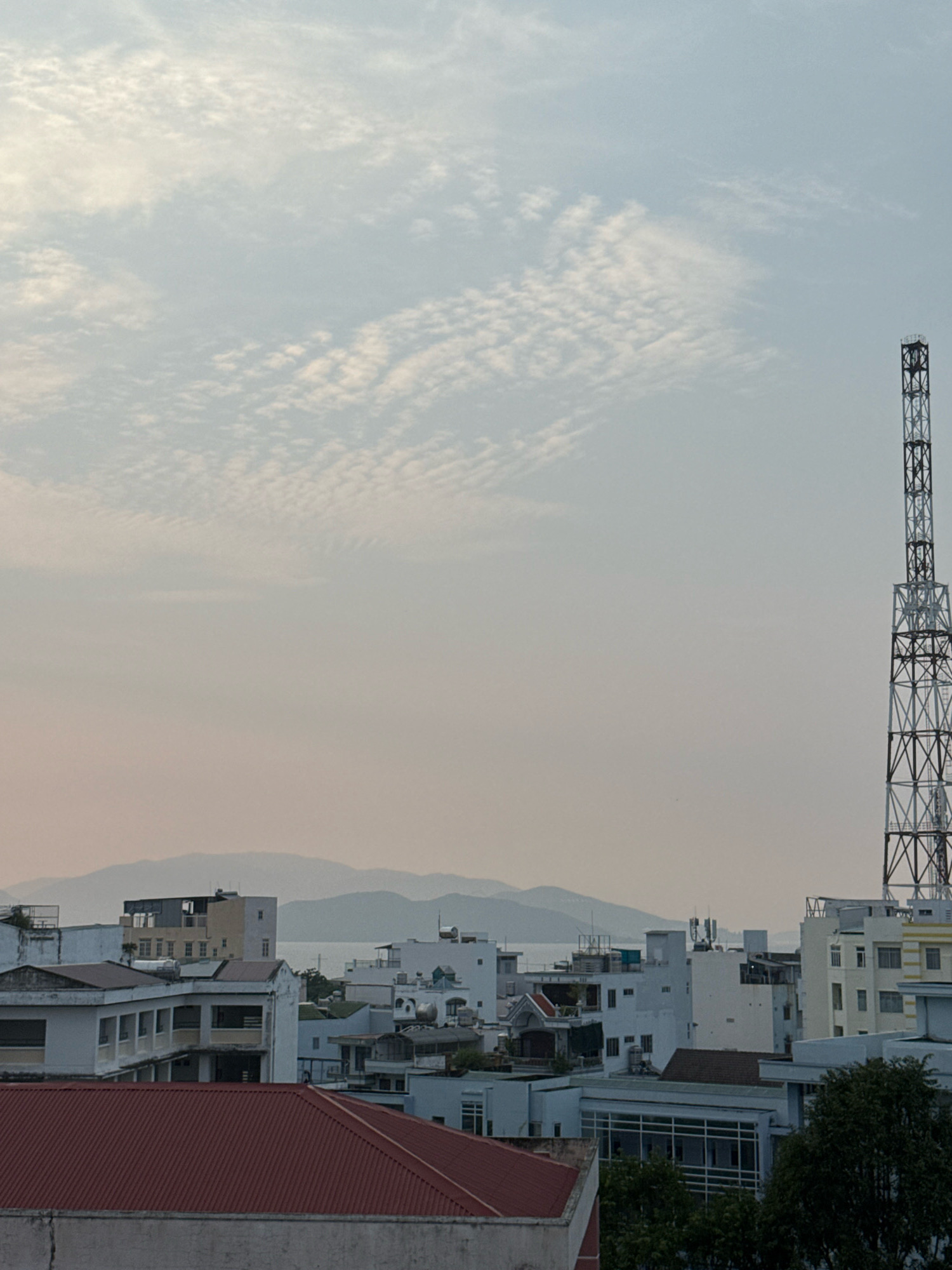 This image depicts an urban landscape in Nha Trang, Vietnam, likely taken during the late afternoon or early evening due to the soft, diffused light and pale sky. The foreground is dominated by the rooftops of several buildings, including a prominent red-tiled roof in the lower left quadrant. Beyond these, a dense collection of multi-story buildings, characteristic of a city, stretches across the mid-ground. These structures vary in color and architectural style, with some featuring balconies, rooftop units, and differing window designs. In the background, a hazy line of mountains and the sea are visible, suggesting the city's coastal location. To the right, a tall, industrial-style radio tower stands prominently against the sky, a common sight in urban areas. The sky itself is filled with wispy, scattered clouds. No people are visible in the frame. The overall scene is one of a developing urban environment against a natural backdrop.