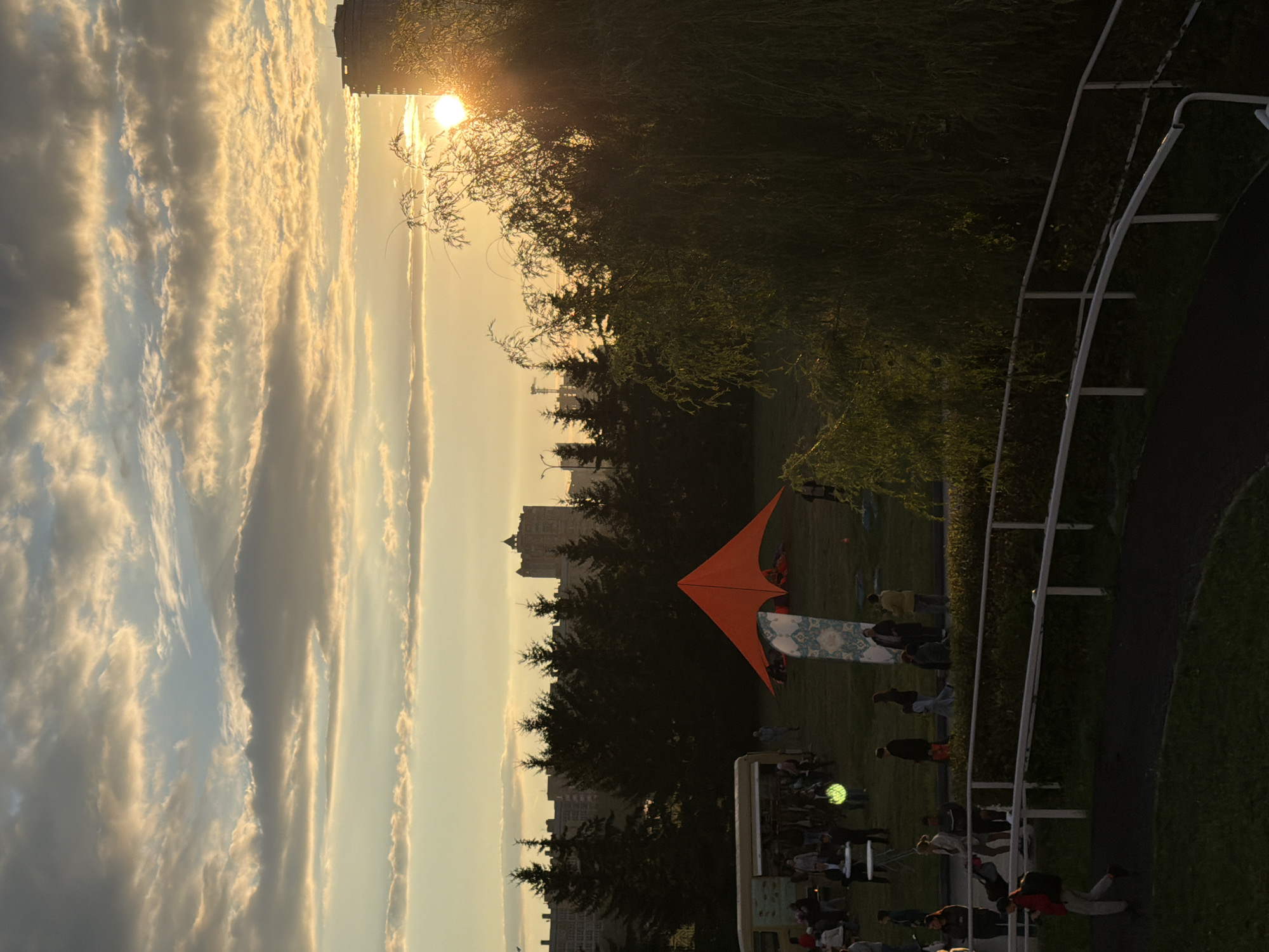 An outdoor event is taking place at dusk. The sky is filled with dramatic clouds, illuminated by the setting sun which is peeking out from behind a tall building on the right. In the foreground, a curved white railing separates a grassy area from a dark pathway. People are scattered across the grassy field, with some gathered around a food truck on the left and others near a bright orange, pyramid-shaped tent in the center. Several evergreen trees form a silhouette against the skyline, behind which are various city buildings. A large, weeping willow tree with long, drooping branches partially obscures the view on the right side of the image. The overall atmosphere is one of a casual gathering as the day ends.