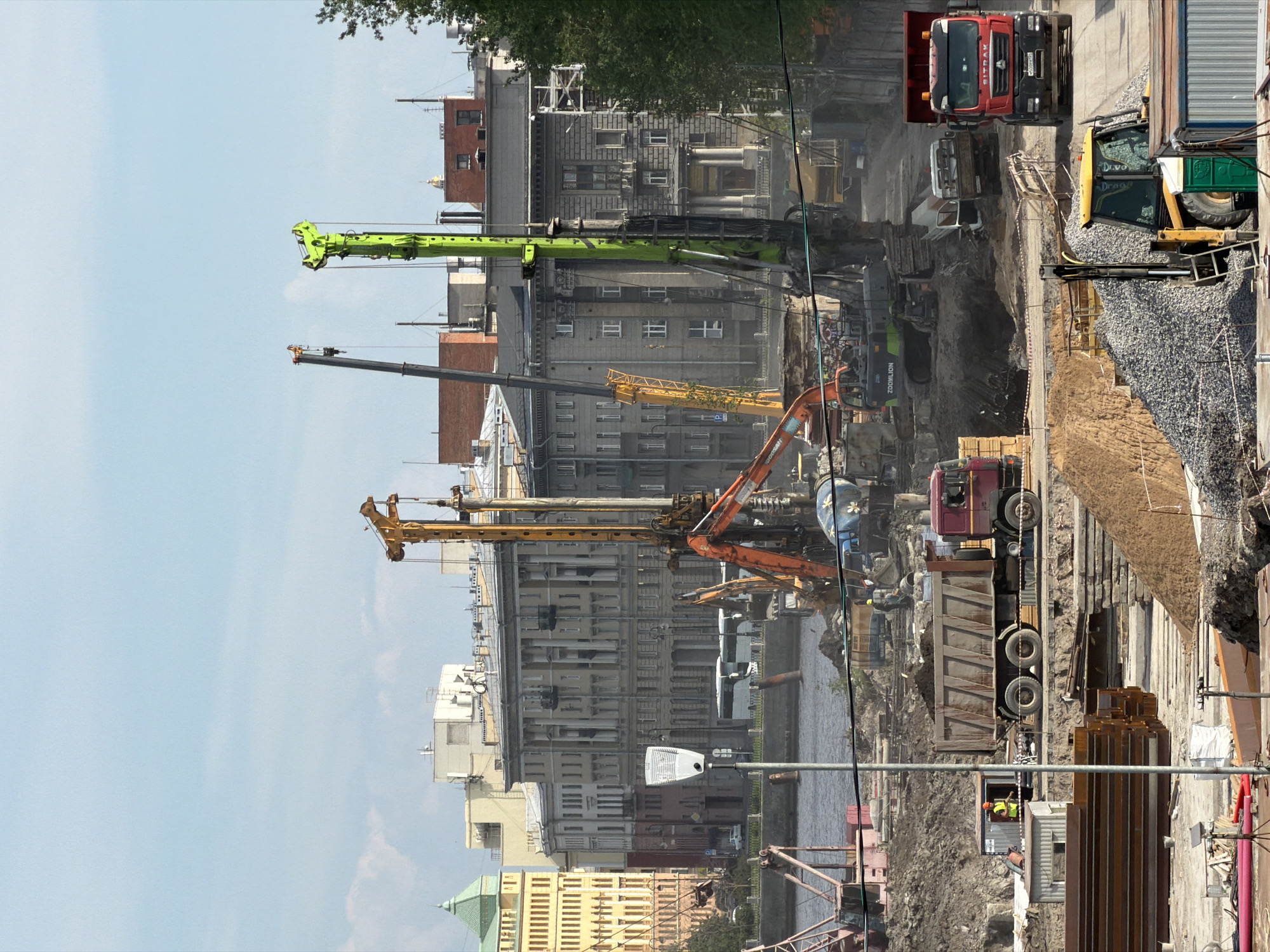 This image depicts an active large-scale construction site in Saint Petersburg, Russia, situated along a wide river. The scene is observed during daytime under a partly cloudy, light blue sky. In the foreground and midground, a significant area of disturbed earth constitutes the construction zone. This area features exposed soil, mounds of sand, and piles of grey aggregate. Numerous pieces of heavy machinery are actively present or positioned for work. Three prominent piling rigs/drilling machines are visible: one dark yellow/orange, one dark grey, and one bright lime green. The green rig, branded "ZOOMLION," is particularly tall and equipped for deep foundation work. A green "ZOOMLION" tracked excavator is positioned near the base of one of the rigs. At least three dump trucks are visible. A maroon semi-trailer dump truck, loaded with earth, is parked on a dirt track in the lower-left midground. To its right, a red "SITRAK" dump truck with a visible Russian license plate (M500PP193) is moving towards the viewer. A smaller white flatbed truck is positioned behind the SITRAK truck, adjacent to the green ZOOMLION excavator. In the lower right foreground, a yellow backhoe loader is partially visible, obscured by a green portable toilet. A blue concrete mixer truck, featuring white and blue painted details, is located in the mid-left. Construction materials are scattered throughout the site, including stacks of reddish-brown metal beams/sheet piling, red pipes, and various other components. Several portable cabins or containers are present; one white cabin with a yellow door is seen in the lower-left, and another grey structure is nearby. Two individuals in yellow high-visibility vests and hard hats are discernible. One stands near the white portable cabin, next to the maroon dump truck. Another is observed near the blue concrete mixer truck. A tall, grey street light pole with a white, modern fixture stands prominently in the lower-left center, separating portions of the construction site. A wide body of water, likely the Neva River or a substantial canal, spans the midground, separating the construction site from the urban background. Some dark, vertical structures, possibly old piles or foundations, protrude from the water near the construction edge. A white passenger boat or river bus is visible on the water, moving towards the left. The background reveals a cityscape characteristic of Saint Petersburg. On the far left, a distinctive multi-story yellow building with a green conical roof or spire is prominent. Along the