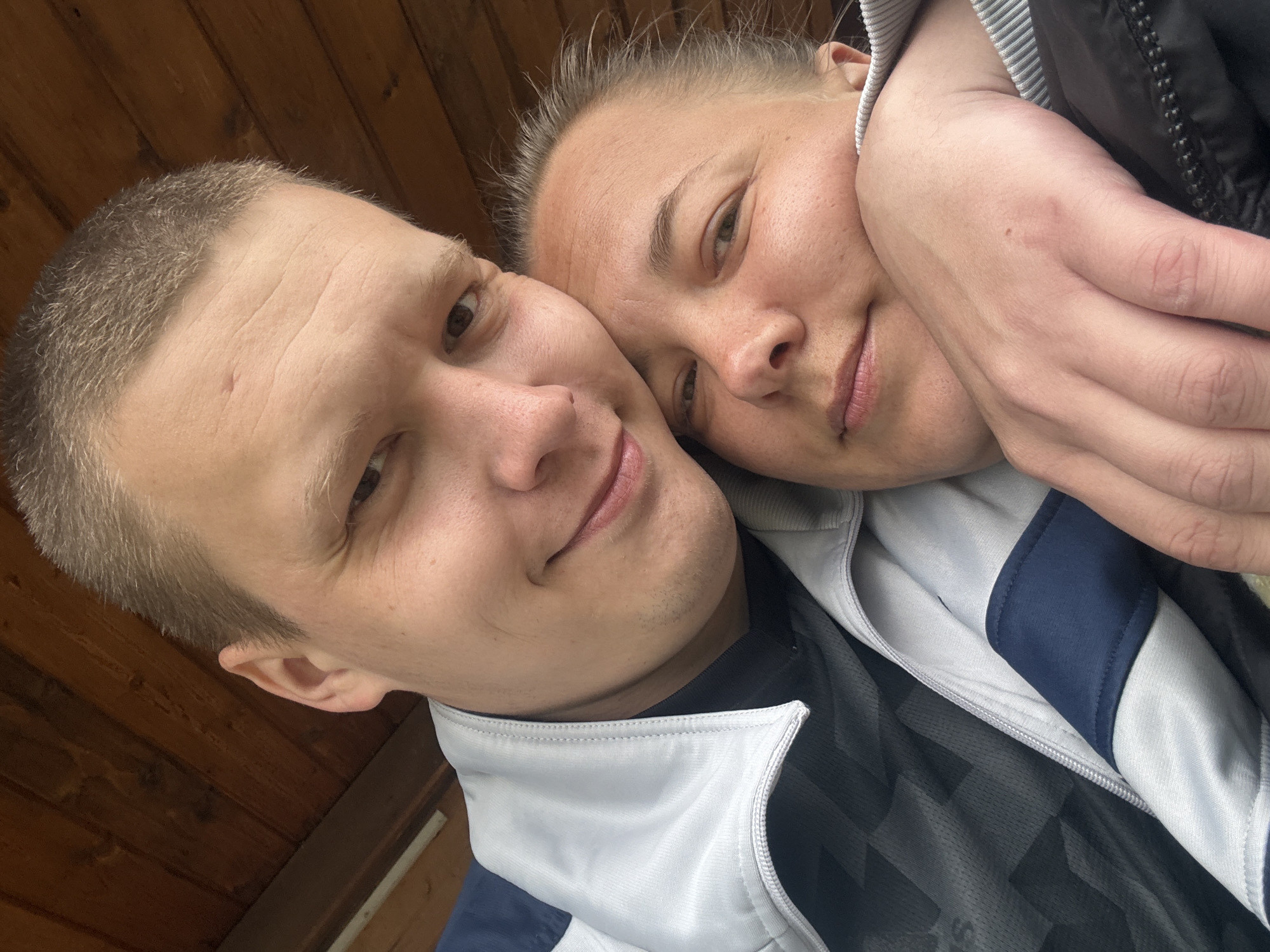 The image depicts a close-up selfie of a man and a woman. The man has short, light-colored hair and is wearing a gray and blue zip-up jacket over a black shirt. The woman has her hair pulled back and is leaning her head against the man's. Her hand is visible near her face. The background appears to be a wooden wall. The location is stated to be Dmitrov, Russia.