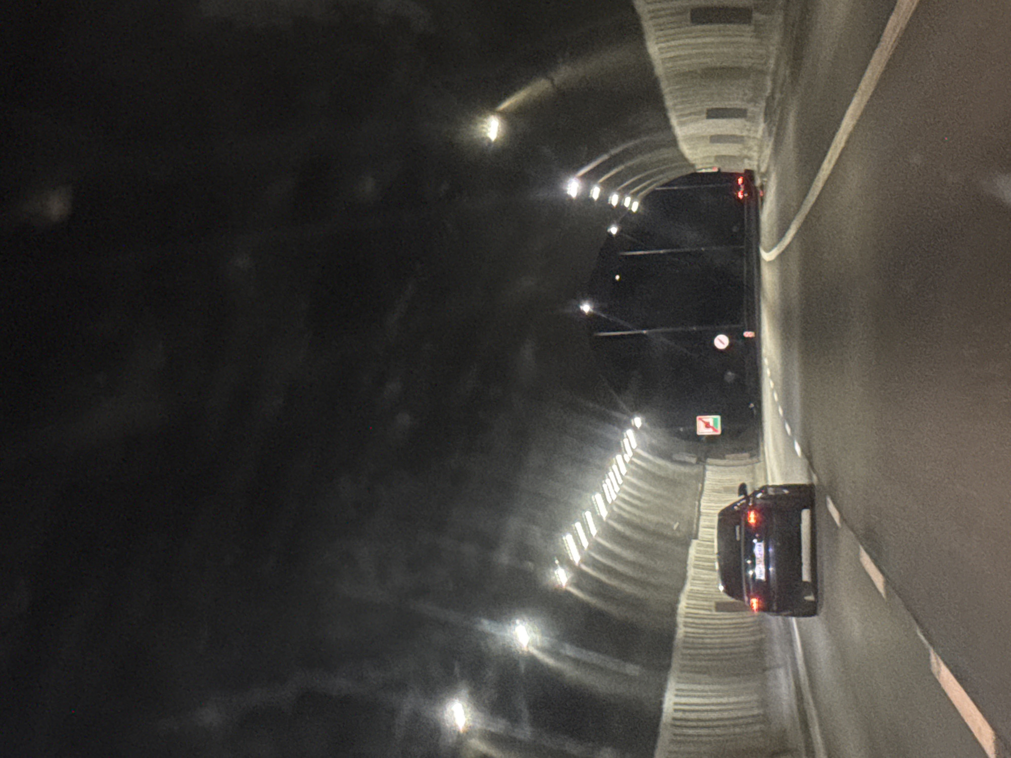 The image depicts the inside of a tunnel, likely a road tunnel, with two vehicles visible. The tunnel walls are curved and lined with evenly spaced lights. The vehicle closer to the viewer is a dark-colored passenger car, displaying its rear lights. Further inside the tunnel is a larger, dark vehicle that could be a bus or van. A sign with a green border is visible above the second vehicle. The road surface is light gray with a lane divider line. Based on the location information, the tunnel is located in or near Dolna Vasilitsa, Bulgaria.