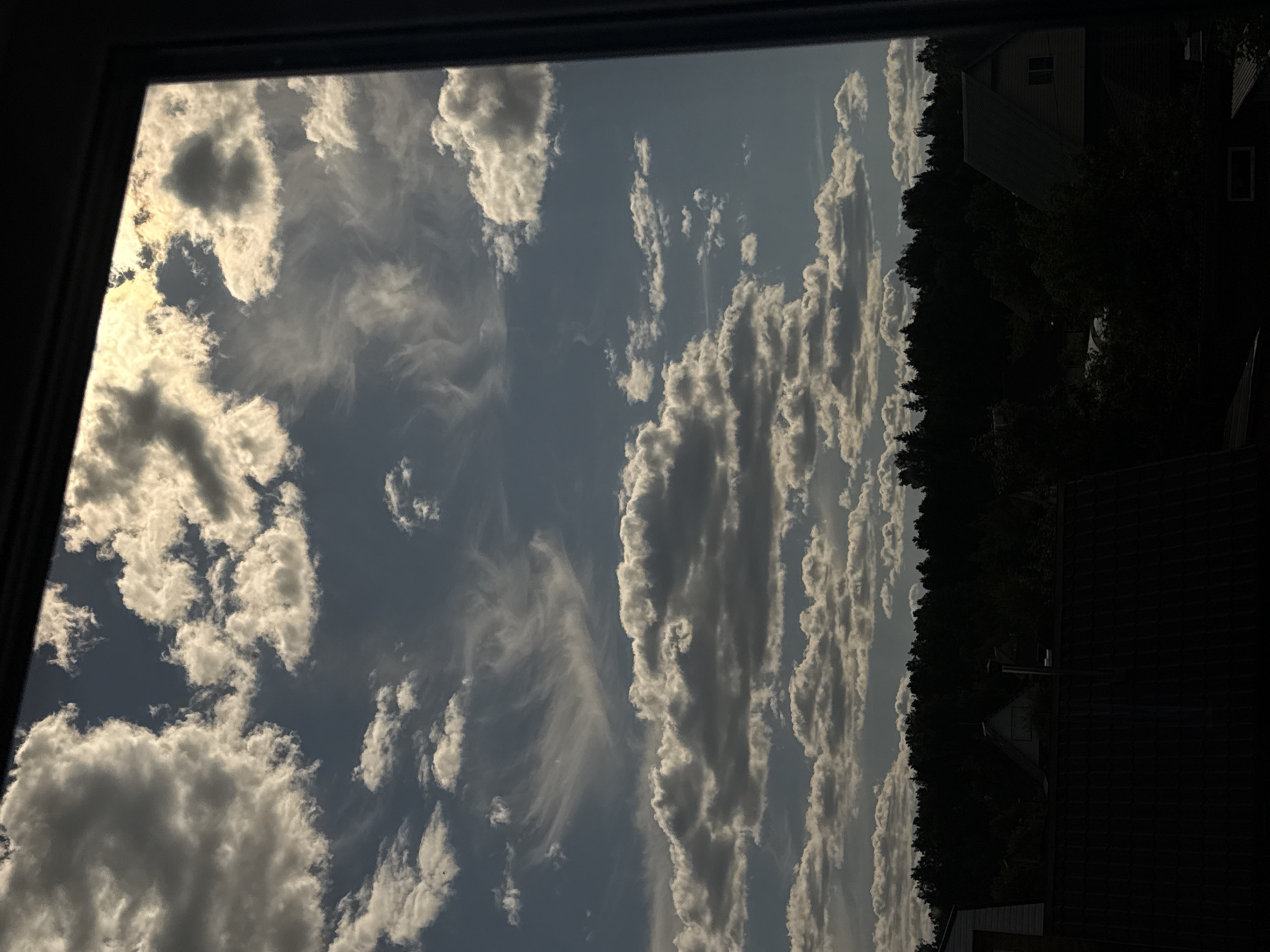 The image depicts a static outdoor scene viewed through a dark window frame, allegedly located in Lenino, Russia. The upper two-thirds of the vertical composition are dominated by a partly cloudy sky against a medium blue-grey backdrop. The sun, not directly visible, illuminates numerous cumulus and stratocumulus clouds with bright white and yellow highlights, particularly in the upper right quadrant, suggesting a strong backlighting effect. Wispy cirrus clouds are also present in the higher atmosphere. A dark, dense treeline stretches horizontally across the lower portion of the sky, casting the trees into silhouette. Below the treeline, several residential structures are visible, mostly in deep shadow; these include a white gable end on the far left and an angled, dark-roofed house with a lighter wall towards the center-right. The immediate foreground, at the bottom of the image, is entirely obscured by deep shadow, likely the viewer's roof or windowsill. There are no visible people, animals, or discernible actions.