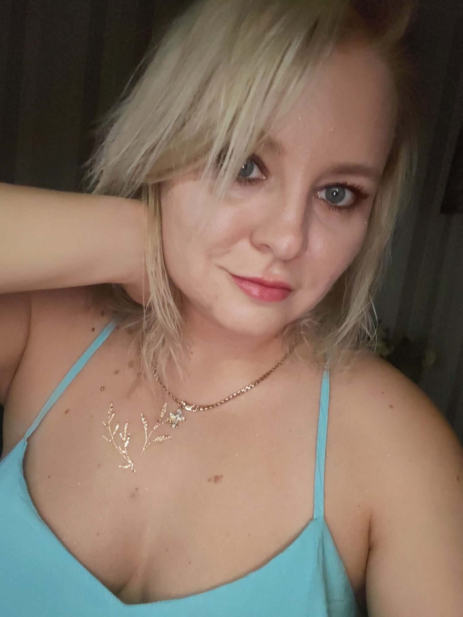 The image shows a close-up selfie of a blonde woman with blue eyes and pink lipstick, wearing a blue top and a gold necklace. There is a light-colored glittery design on her chest. The background is dark and blurry, with hints of dark curtains or walls. There is no indication of the location being in Izhevsk, Russia.