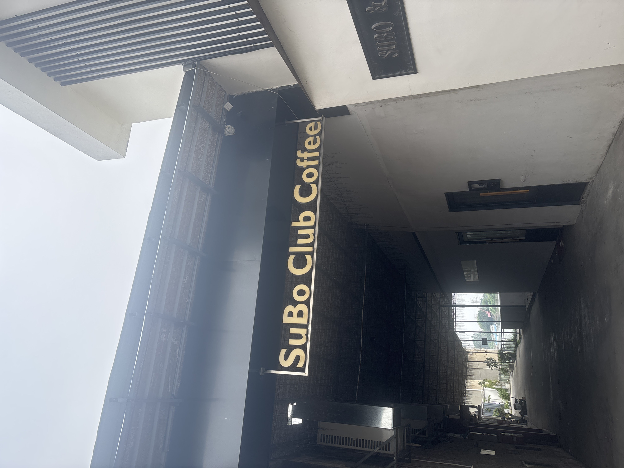 This image depicts the exterior of a commercial establishment in the city of Nanhai District, China, identified by a prominent sign that reads "SuBo Club Coffee." The sign is illuminated and hangs above an entryway to what appears to be a covered walkway or alley. The architecture is modern, with a combination of white walls, dark metal accents, and corrugated roofing. Adjacent to the main sign, another smaller, darker sign is visible on a white wall, partially obscured but appearing to also bear the establishment's name. The walkway is dimly lit and shows some signs of industrial elements, such as air conditioning units or ventilation equipment mounted on the walls. Plants in pots are visible further down the walkway, suggesting an attempt at landscaping. The overall scene is overcast, indicating potentially cloudy weather. There are no people visible in the immediate vicinity of the establishment's entrance.