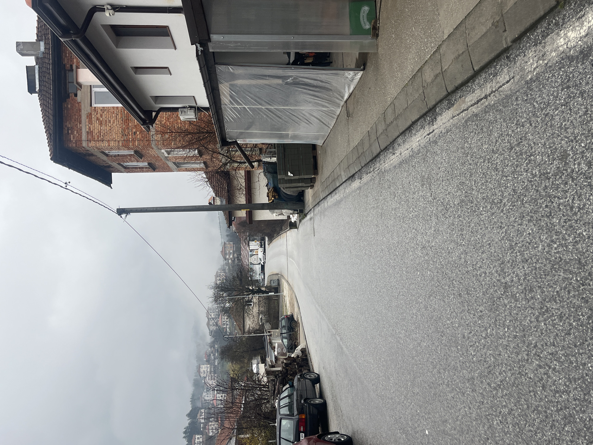 The image depicts a street scene in Srednogortsi, Bulgaria. The view is looking downhill on a wet, grey day, likely after or during rain. A two-story building with a brick facade and a white upper level is on the left side of the street. The lower part of the building appears to be undergoing renovation or construction. A covered outdoor storage area abuts the building. Cars are parked along the right side of the street, facing downhill. Utility poles and wires run alongside the street. In the distance, there is a hillside covered with buildings and trees, partially obscured by mist or fog.