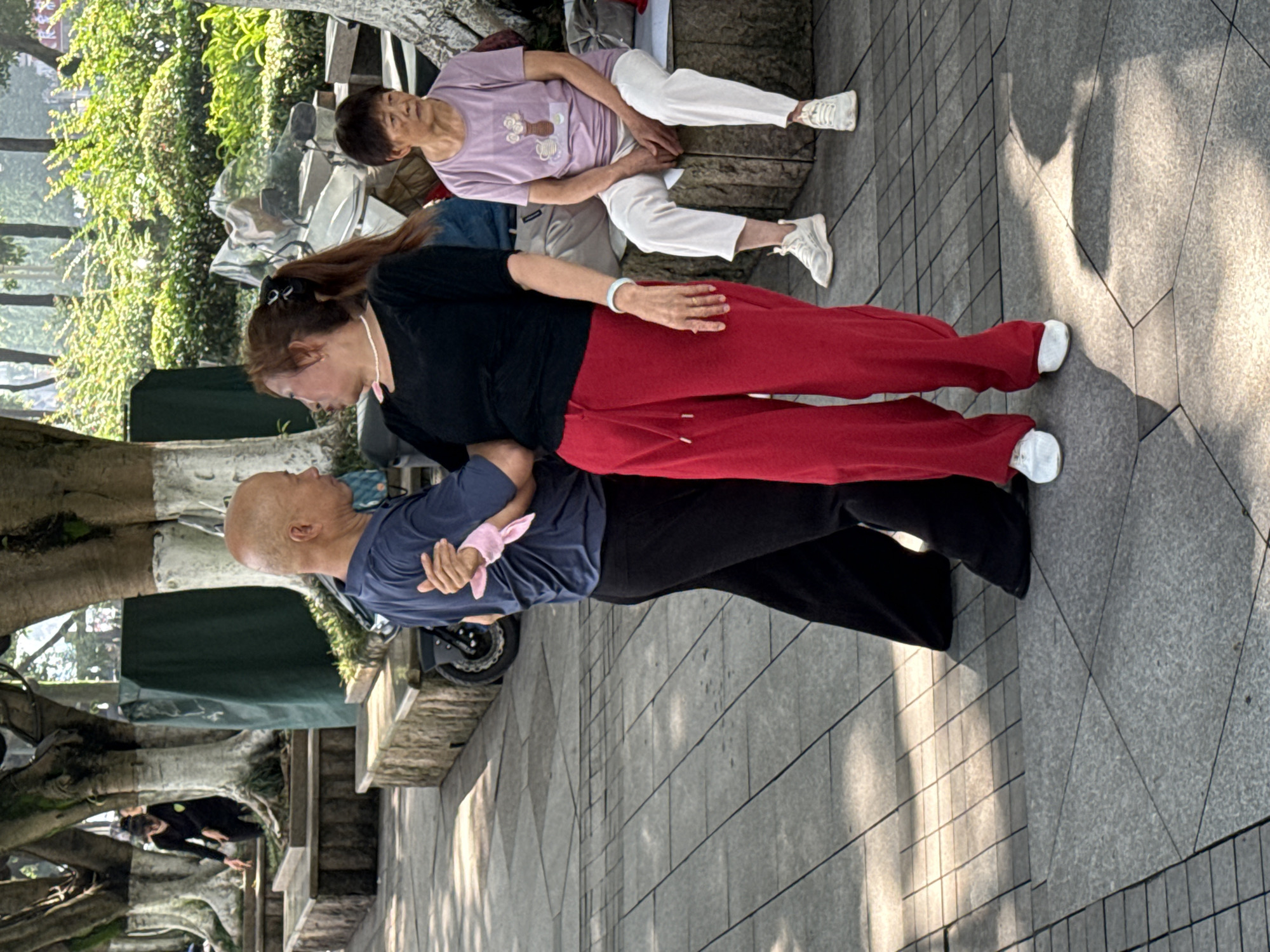In Jiangjin, China, an elderly couple dances outdoors on a paved area lined with trees. The man, bald and wearing a navy blue t-shirt and black pants, holds his partner's hand. His left hand is wrapped in a pink fabric. His partner, with her hair pulled back in a ponytail and wearing a black t-shirt and vibrant red pants, gazes at him. Behind them, an elderly woman sits on a stone bench, wearing a light purple t-shirt and white pants. Another person in black clothing is visible in the background, bending down near a motorcycle. The scene is bathed in sunlight, casting shadows on the ground.