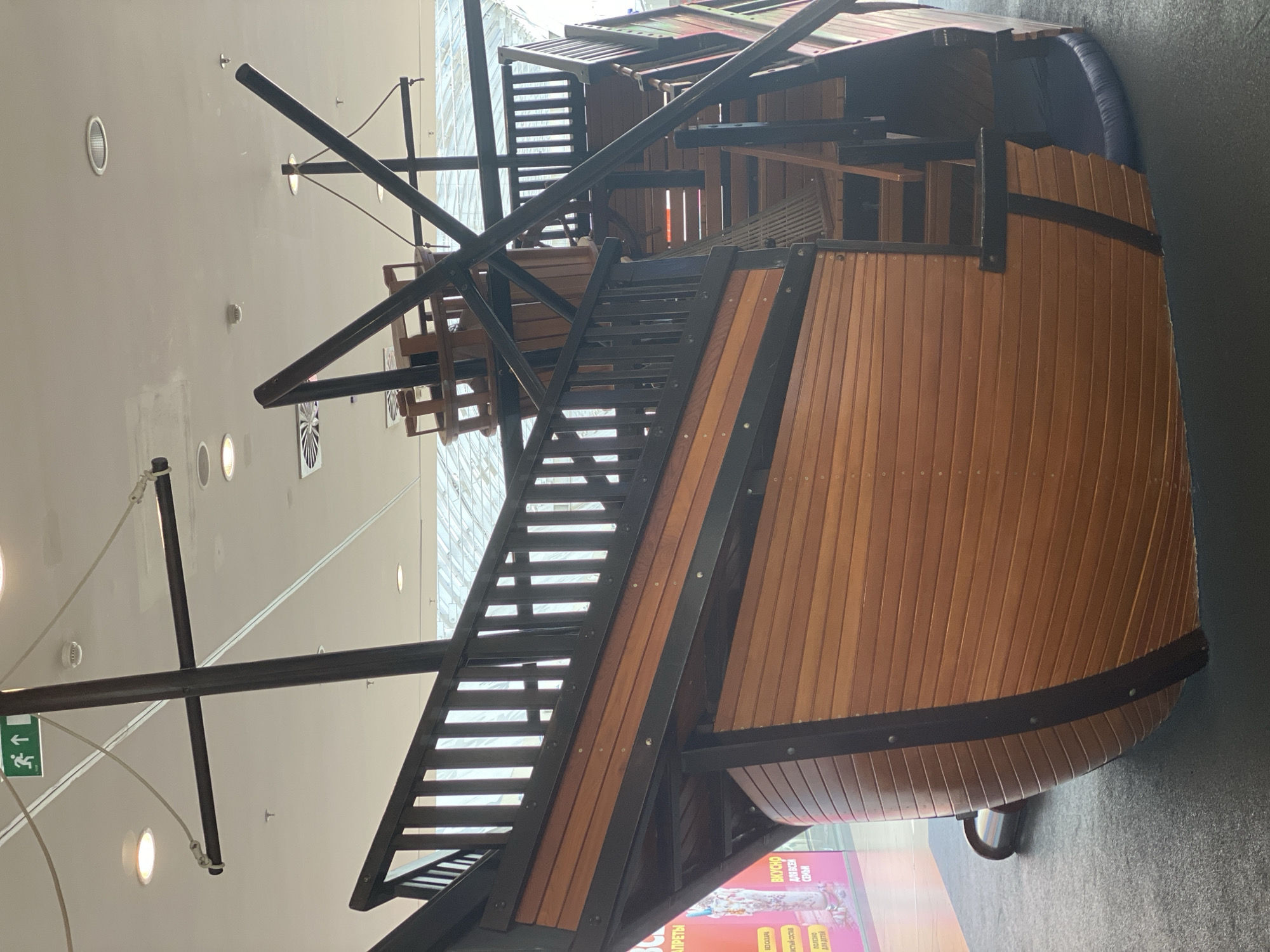 The image shows a wooden play structure designed to resemble a pirate ship. The ship is brown and black, with a detailed wooden hull and railings. Stairs are visible leading up to the deck of the ship. The play structure is located indoors, on a dark gray carpeted floor, under a white ceiling with fluorescent lights. In the background, a large glass ceiling or window allows natural light to enter, revealing a glimpse of the sky and possibly other parts of the building. A green emergency exit sign with arrows is visible on the ceiling. A blurred advertisement is partially visible to the left of the ship. The scene appears to be a children's play area within a shopping mall or similar public indoor space. The location in Novokuznetsk, Russia is not discernible from the visual information provided in the image.