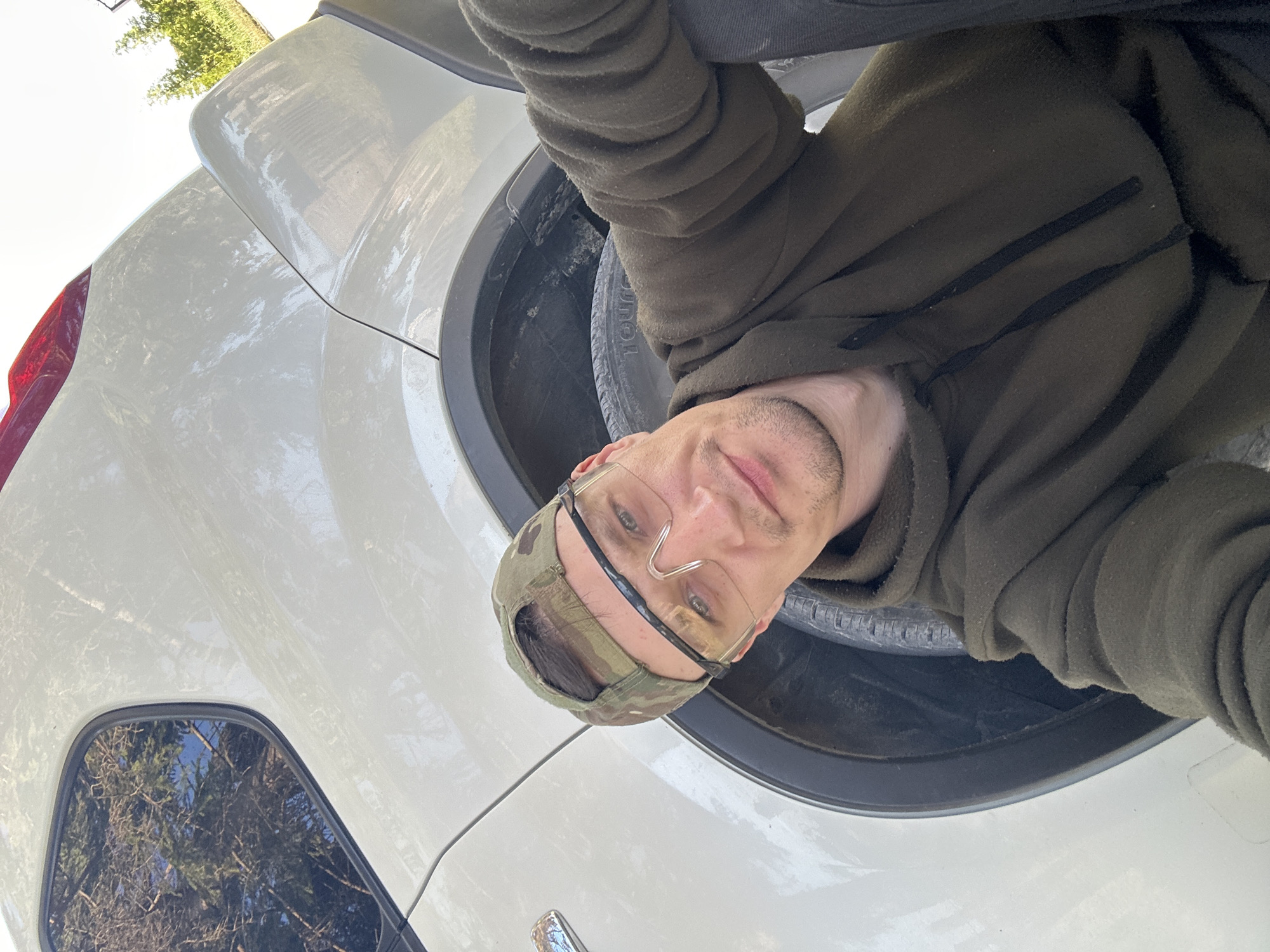 The image shows a selfie of a man in Makiv, Ukraine. He is wearing a camouflage baseball cap and safety glasses, and a dark green hooded sweatshirt. He is positioned in front of the wheel well of a white vehicle. The vehicle appears to be parked outdoors, with trees and a bright sky visible in the background. The man is looking directly at the camera, with a slight smile.