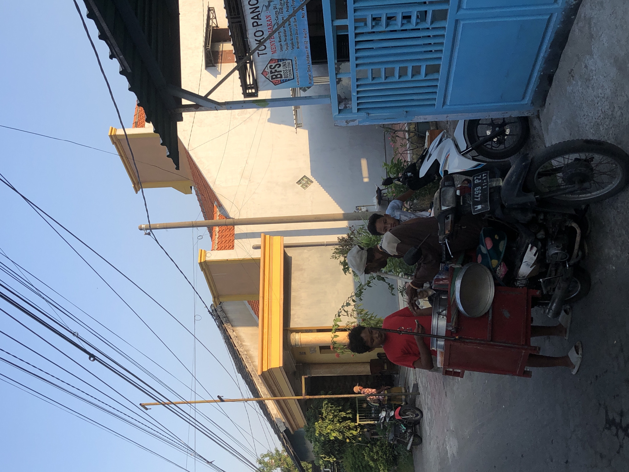 In Kalidawir, Indonesia, two young males and an older male are engaged in a transaction near a street. A young male, wearing a red t-shirt and light-colored slippers, stands to the left, appearing to be the vendor, with a food cart attached to a motorcycle. The cart, a red wooden structure, holds metal containers, one of which is covered by a lid. An older male, in a brown and tan shirt and a light-colored cap, is on the right, reaching towards the cart, possibly making a purchase or receiving food. Another young male in a light-colored shirt is partially visible behind the older male, observing the interaction. Two motorcycles are parked nearby: a black motorcycle in the foreground with a visible license plate "W 4499 PZ 11-17", and a white motorcycle behind it. To the right, a bright blue metal gate is prominent, and a shop sign above it reads "TOKO PANCA" and "BFS BOSS UNA FISHING STORE". The background features houses with orange tiled roofs and yellow decorative elements, and overhead, a tangle of electrical wires stretches across the clear blue sky. Trees and foliage are visible on the left side of the scene. The overall atmosphere suggests a typical street scene during daylight hours.