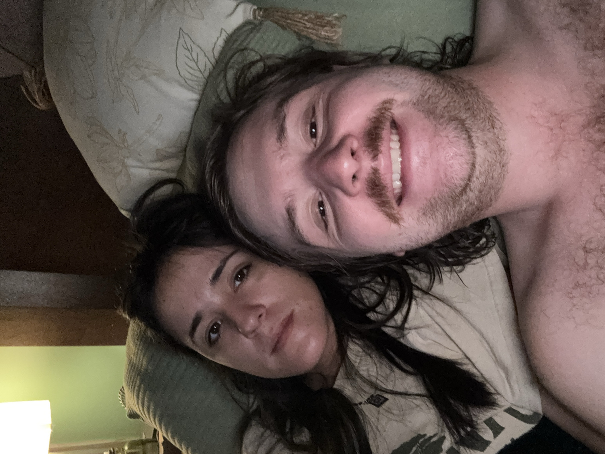 A close-up, slightly rotated indoor shot depicts a man and a woman lying down next to each other, presumably in a bed. The man is on the right, smiling with a mustache, and has long hair. He is shirtless and fair-skinned. The woman is on the left with long, dark hair. They are lying on pillows and bedding. A lamp is visible in the bottom left corner. There is no information in the image to suggest the location is Huixquilucan de Degollado, Mexico.
