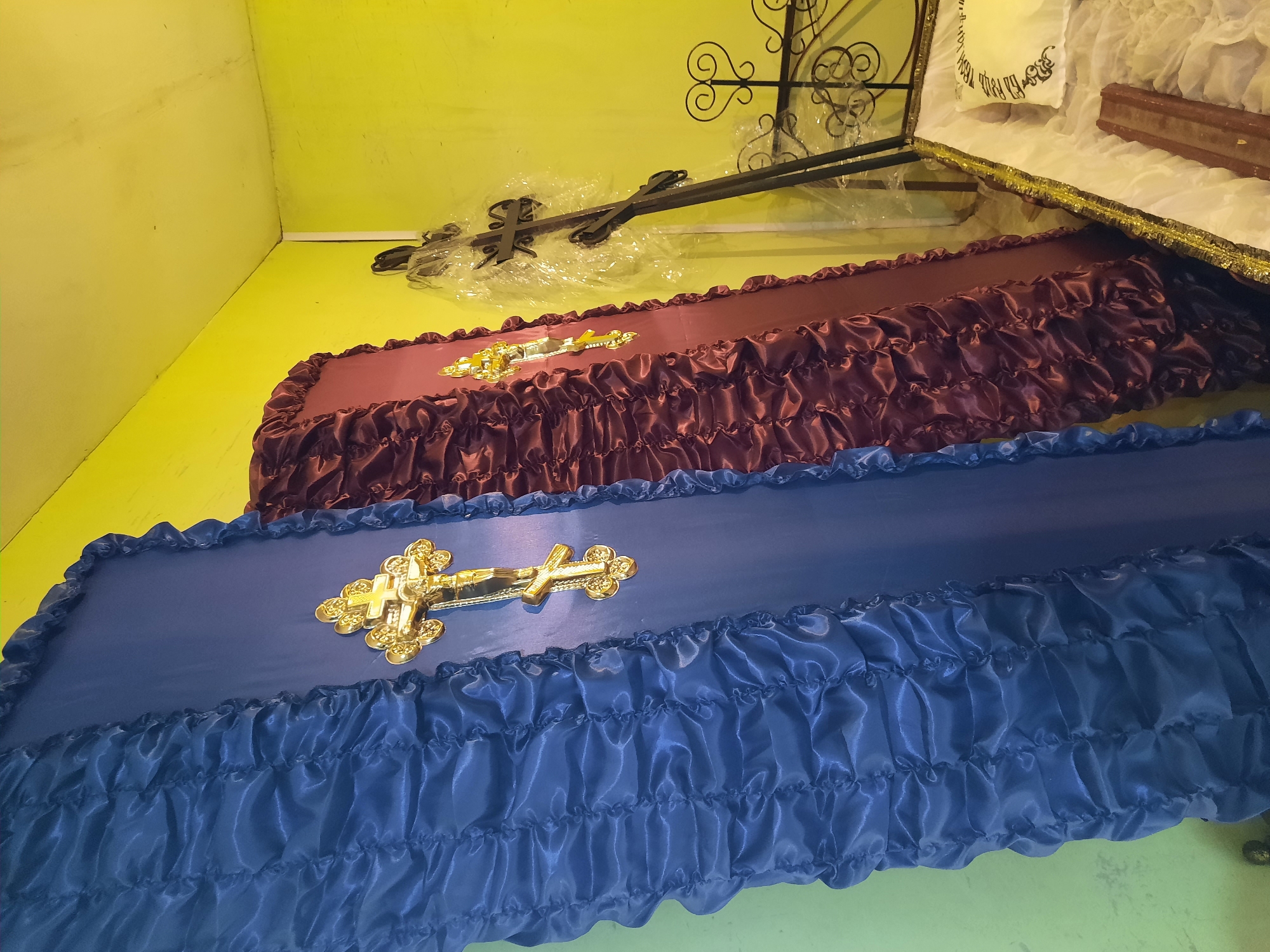 The image shows a room in Krasnogorskoye, Russia, with yellow walls and coffins. Two coffins, one blue and one burgundy, are prominently displayed with golden crosses on top. Wrapped crosses are resting on the wall. Another coffin, partially visible, is open and lined with white fabric, featuring decorative elements.