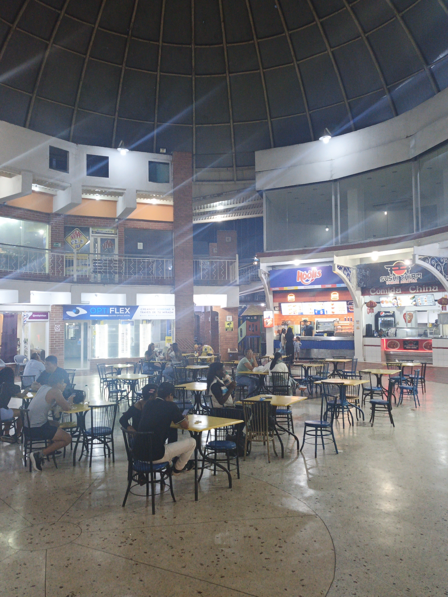 This image captures a bustling indoor food court or commercial area, likely within a shopping center, in Tinaquillo, Venezuela. The expansive space is characterized by a prominent domed ceiling with a grid-like structure, suggesting a modern architectural design.
On the ground floor, numerous people are seated at tables with yellow tops and dark chairs, engaging in dining and social interaction. Many individuals are casually dressed, appearing relaxed as they eat or converse. On the right side, two food stalls are visible: "Hoolis," offering fast food, and "Sushi Thiam Comida China," specializing in Asian cuisine, including sushi. People are also seen ordering from these stalls.
The left side of the image reveals a multi-level structure with additional storefronts. A notable business on the ground level is "OPTIFLEX CENTRO ÓPTICO DIGITAL," an optician, with the tagline "CREANDO SONRISAS A TRAVÉS DE TU MIRADA" (Creating smiles through your gaze). An upper-level store displays a "Bienvenidos" sign and a "VIVE 93.8 FM" logo. The flooring is a light-colored terrazzo material with dark flecks.
The scene appears to be taking place in the evening or at night, as the space is illuminated by artificial lighting, creating bright reflections and some lens flares from overhead fixtures. The overall atmosphere is one of casual leisure and daily activity in a commercial setting.