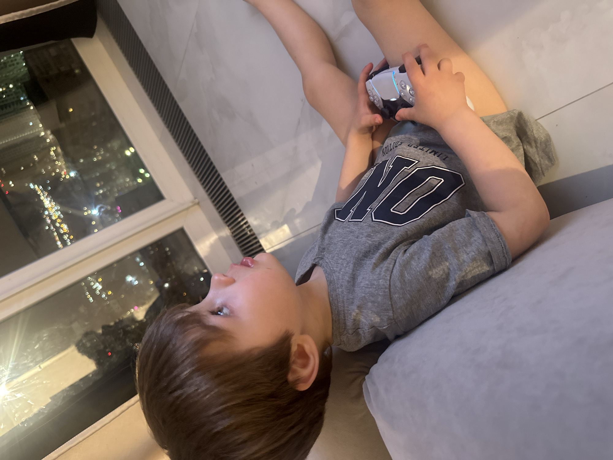 A young boy lies on a light gray sofa, holding a white and silver electronic device, possibly a toy or game controller. He is wearing a gray t-shirt with a dark blue design. To his left is a large window showing a blurred nighttime city scene with numerous lights, which may be Khimki, Russia given the query parameters. The floor is light-colored and appears to be made of tile or stone.