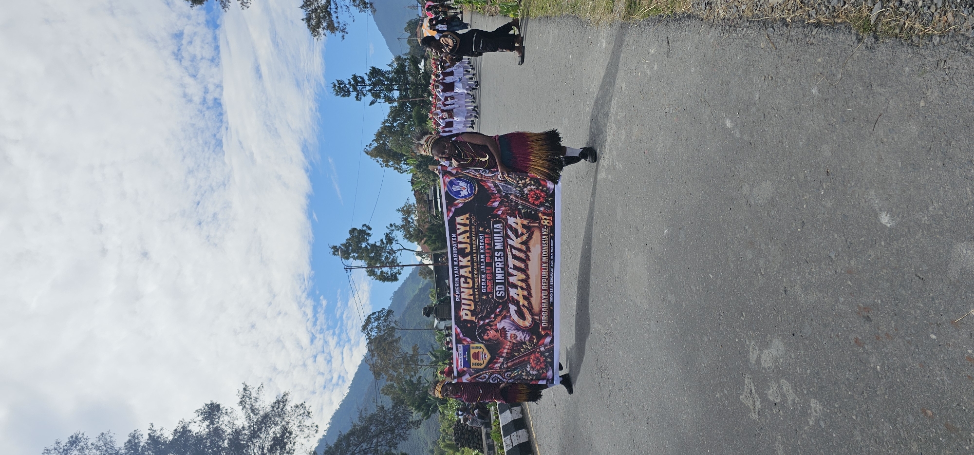 In Puncak Jaya, Indonesia, a parade is taking place on a paved road, with mountains and trees visible in the background under a cloudy sky. The focus of the image is a large banner held by two individuals. The banner features text that reads "PEMERINTAH KABUPATEN PUNCAK JAYA", "DINAS PENDIDIKAN DAN KEBUDAYAAN", "GERAK JALAN KREASI", "REGU PUTRI", "SD INPRES MULIA", and "CANTIKA DIRGAHAYU REPUBLIK INDONESIA KE - 81". The banner also depicts imagery related to Indonesia.
The two individuals holding the banner are dressed in traditional attire. The person on the left appears to be wearing a headdress, a beaded necklace, and a skirt made of tassels. The person on the right is similarly adorned with traditional clothing and accessories. They are walking on the road, holding the banner horizontally.
Behind the banner bearers, a group of children in matching white and red outfits are marching in formation. Further in the background, other participants in the parade and spectators can be seen. The road is flanked by grassy areas and some sparse vegetation. The overall scene suggests a festive community event.