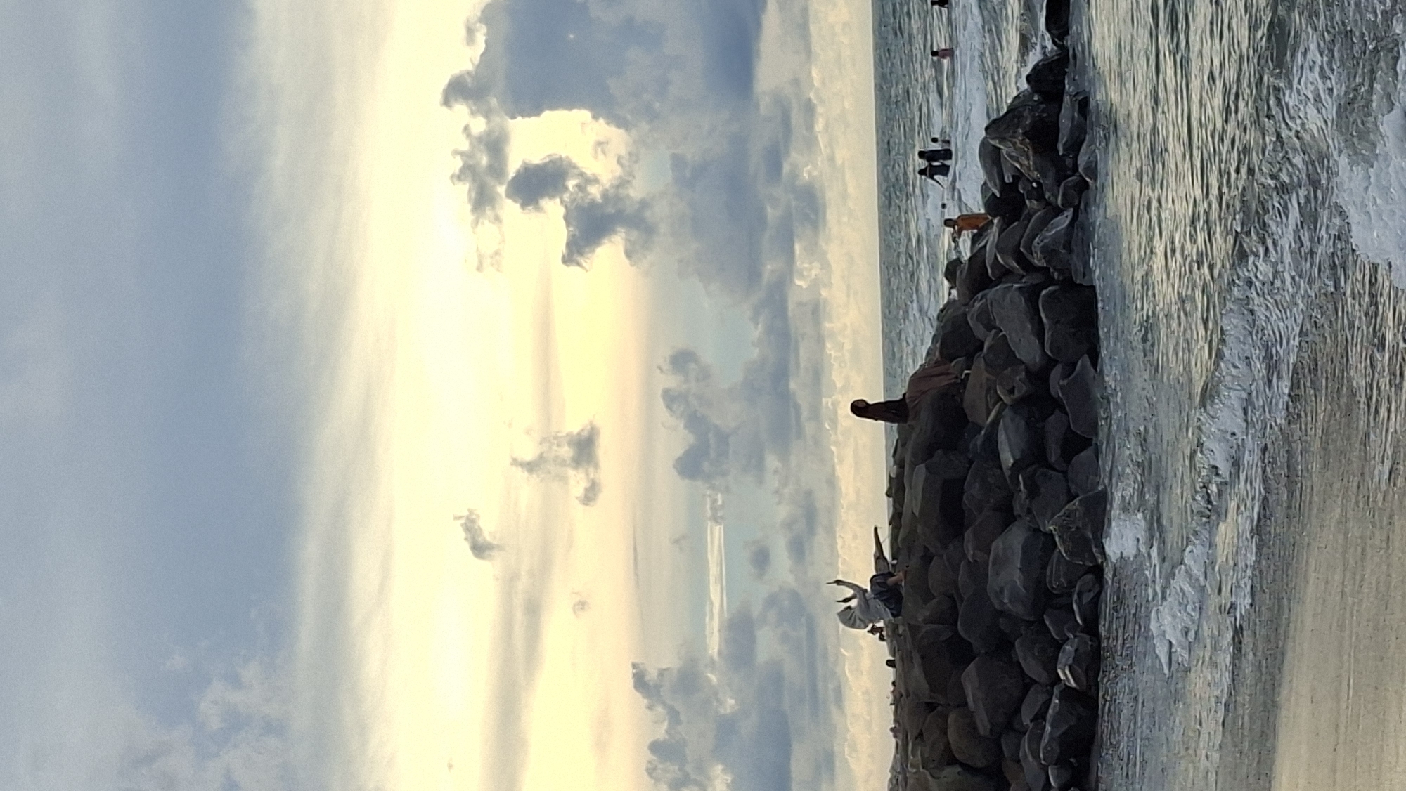 This image captures a sunset scene at Ujong Blang Beach in Lhokseumawe, Indonesia. The sky is a blend of soft blues and yellows, with dramatic, textured clouds. In the foreground, gentle waves lap onto a sandy shore, with the water transitioning to a more turbulent state around a large, dark rock formation that juts into the sea.
Several people are visible on and around the rocks. On the left side of the rock formation, a person in a hijab is seated and appears to be taking a photograph with their phone, holding it up towards the sky. To their right, another person in a hijab is seated on the rocks, facing away from the camera. Further out in the water, several figures are swimming or wading. A person in an orange shirt is standing on the rocks on the right side of the frame, facing the water. The overall atmosphere is serene, with a focus on the natural beauty of the coastline and the activities of beachgoers enjoying the late afternoon light.