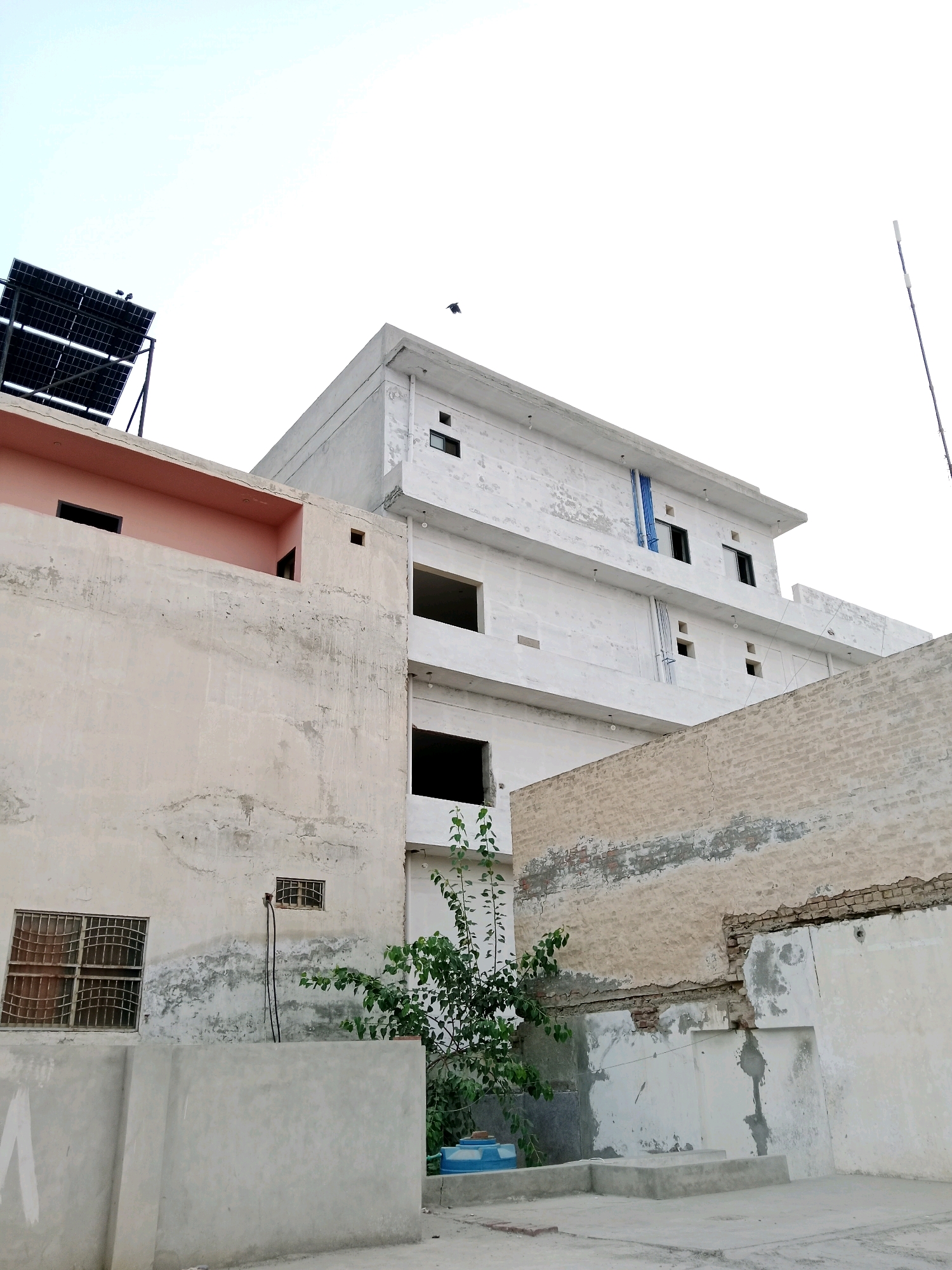 The image captures an exterior view of a multi-story building under construction or renovation, situated in Bahawalpur City Tehsil, Pakistan. The building features a white facade with visible signs of wear and tear, including peeling paint and exposed brickwork. Several windows are present, some are open and black, while others are covered with metal grates. Solar panels are installed on the rooftop of one section of the building. Adjacent to the main structure, there is a lower building with a pink-colored projection and a beige, textured wall with exposed brick. A small tree with green foliage grows near the corner of the beige wall. The sky is overcast with a bright, light-colored haze. The scene appears to be a residential or commercial area, typical of urban environments in Pakistan.
