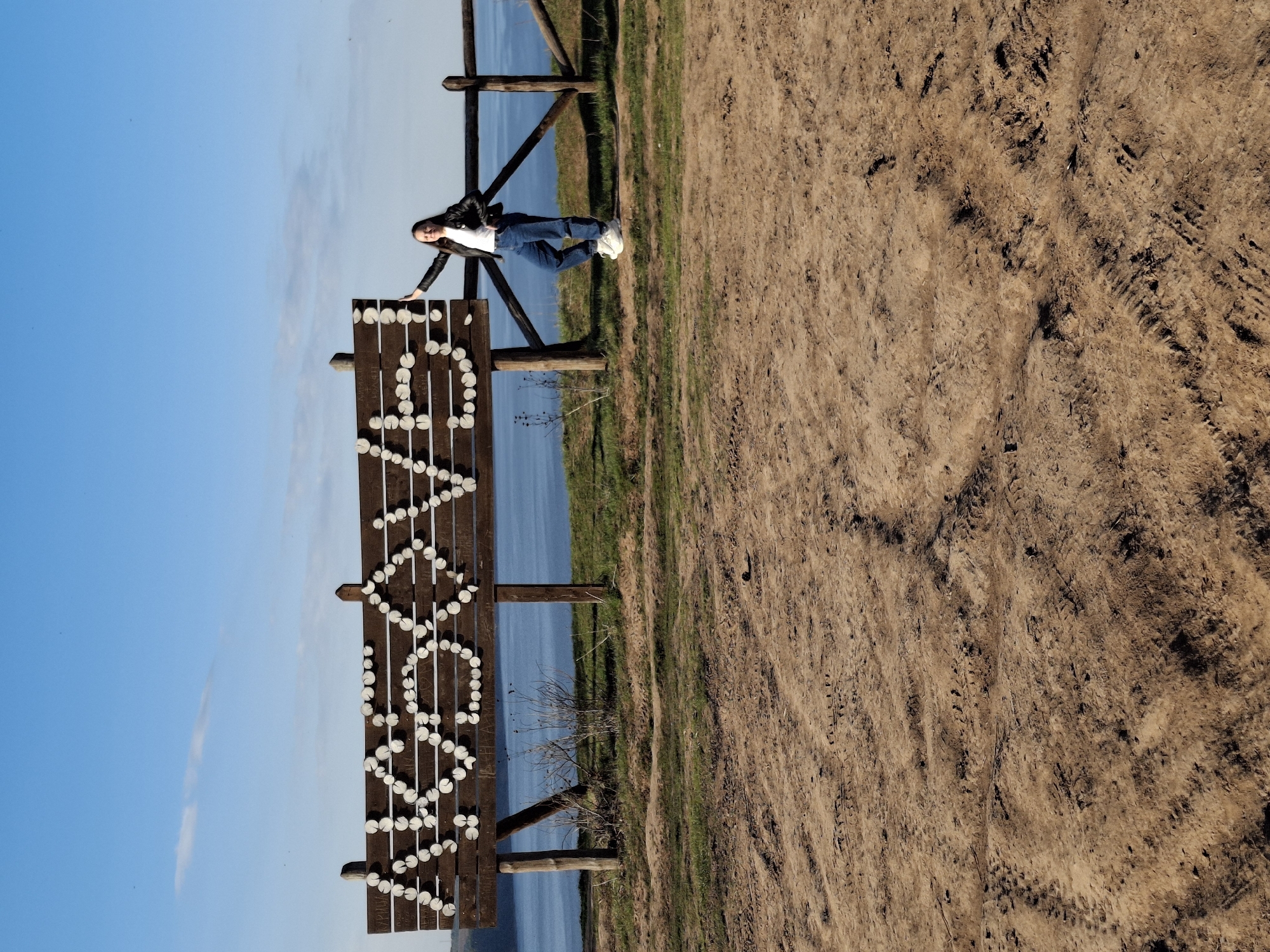 A young woman in blue jeans and a black jacket leans against a wooden sign spelling "VOLODARSK," which is affixed to a wooden fence. Behind the fence is a body of water. The foreground is a dirt and grassy area. The location is Костоваты, Russia.