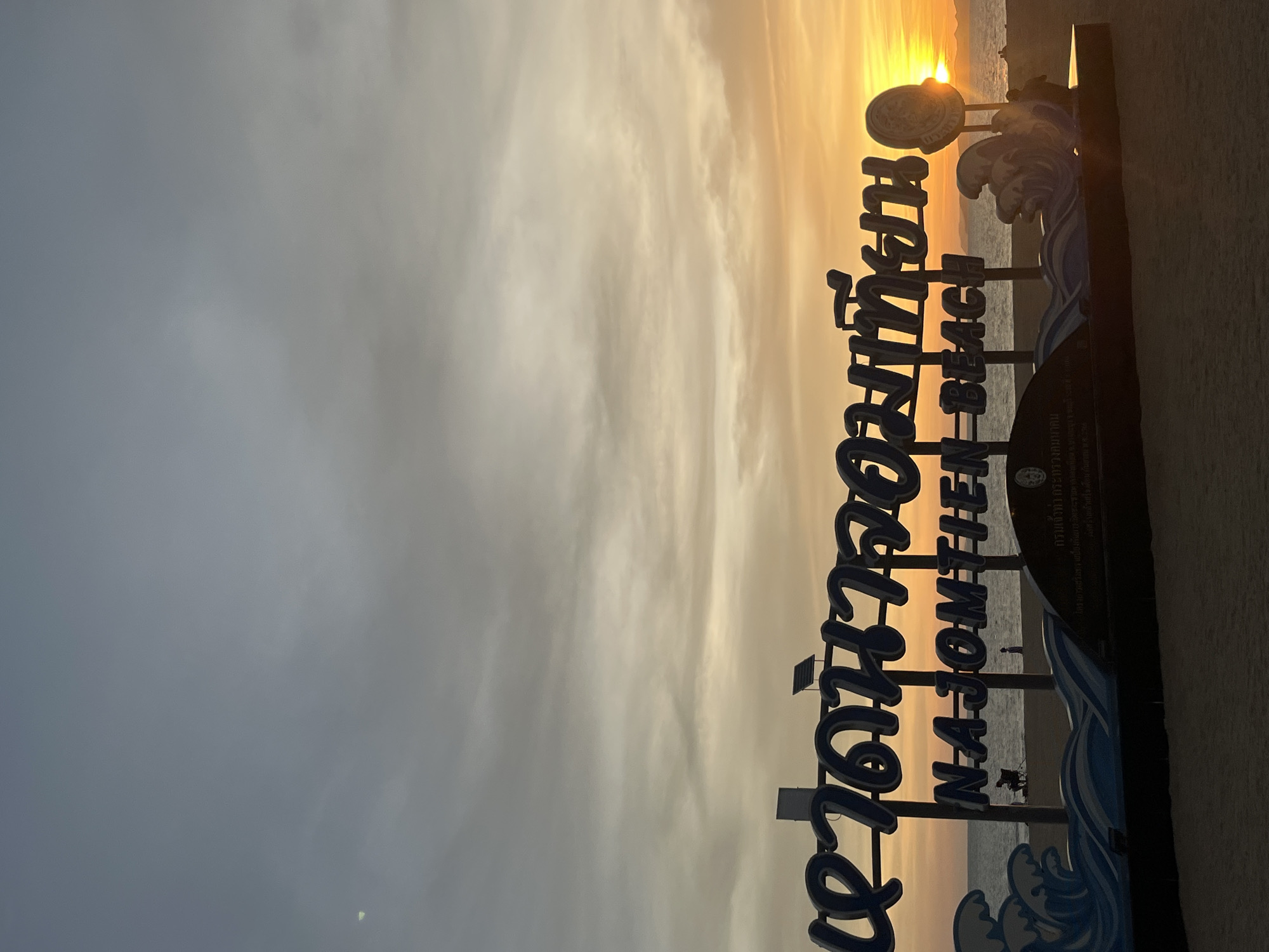 A sign for "Najomthien Beach" is in the foreground. The beach is visible in the background, with a sunset on the horizon. The photo was taken in Pattaya City, Thailand.