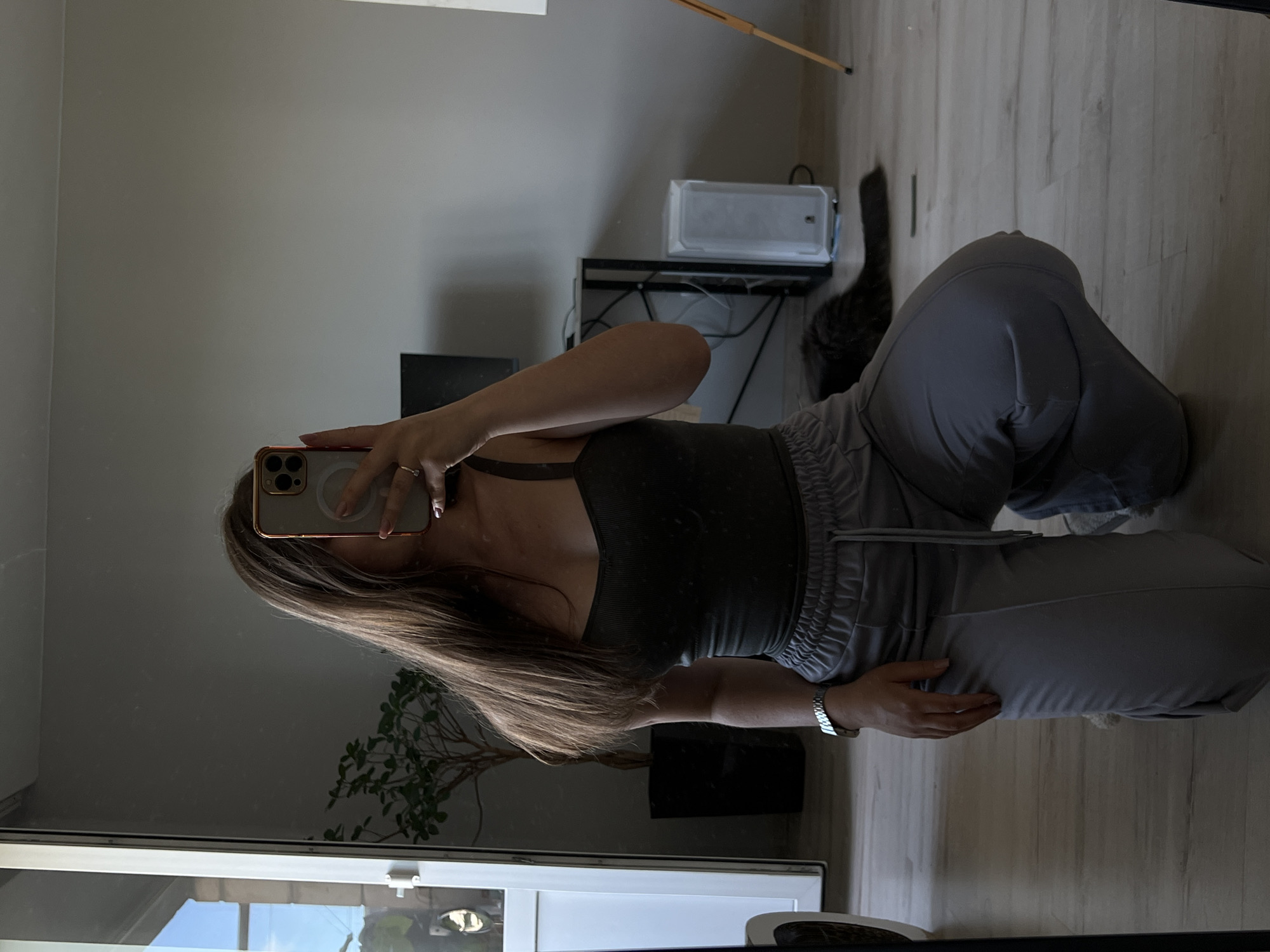 A young woman kneels on a light-colored wood floor, holding a smartphone to take a mirror selfie. She has long, brown hair and is wearing a dark gray tank top and light gray sweatpants. A silver watch is visible on her left wrist. In the background, reflected in a large mirror, is an indoor scene. To the left of the woman is a tall, white door with glass panes, a potted plant, and a black planter. To the right of the woman is a desk with a computer monitor, a white computer tower, and some cables. A dark gray cat is lying on the floor in front of the desk. The room appears to be a residential interior.