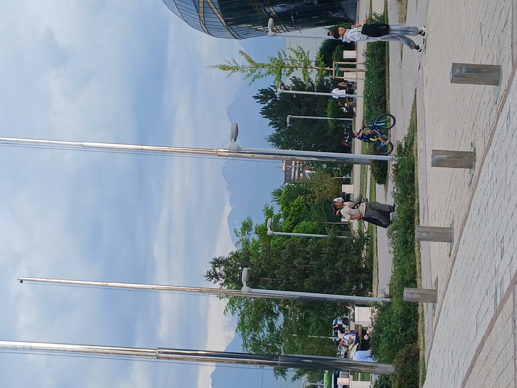 In the city of Unknown, Unknown, an outdoor scene unfolds under a cloudy sky, with mountains in the distance. Tall, silver flagpoles stand prominently in the foreground. A woman walks with a broom and dustpan, seemingly cleaning the paved area. To her left, a young boy rides a blue bicycle on the sidewalk. A group of people are seated on benches in the background, some engaged in conversation, while others appear to be reading. A modern, glass-paneled building with a curved facade is visible on the right side of the image. The ground is paved with light-colored bricks, and there is lush green foliage and trees scattered throughout the area.
