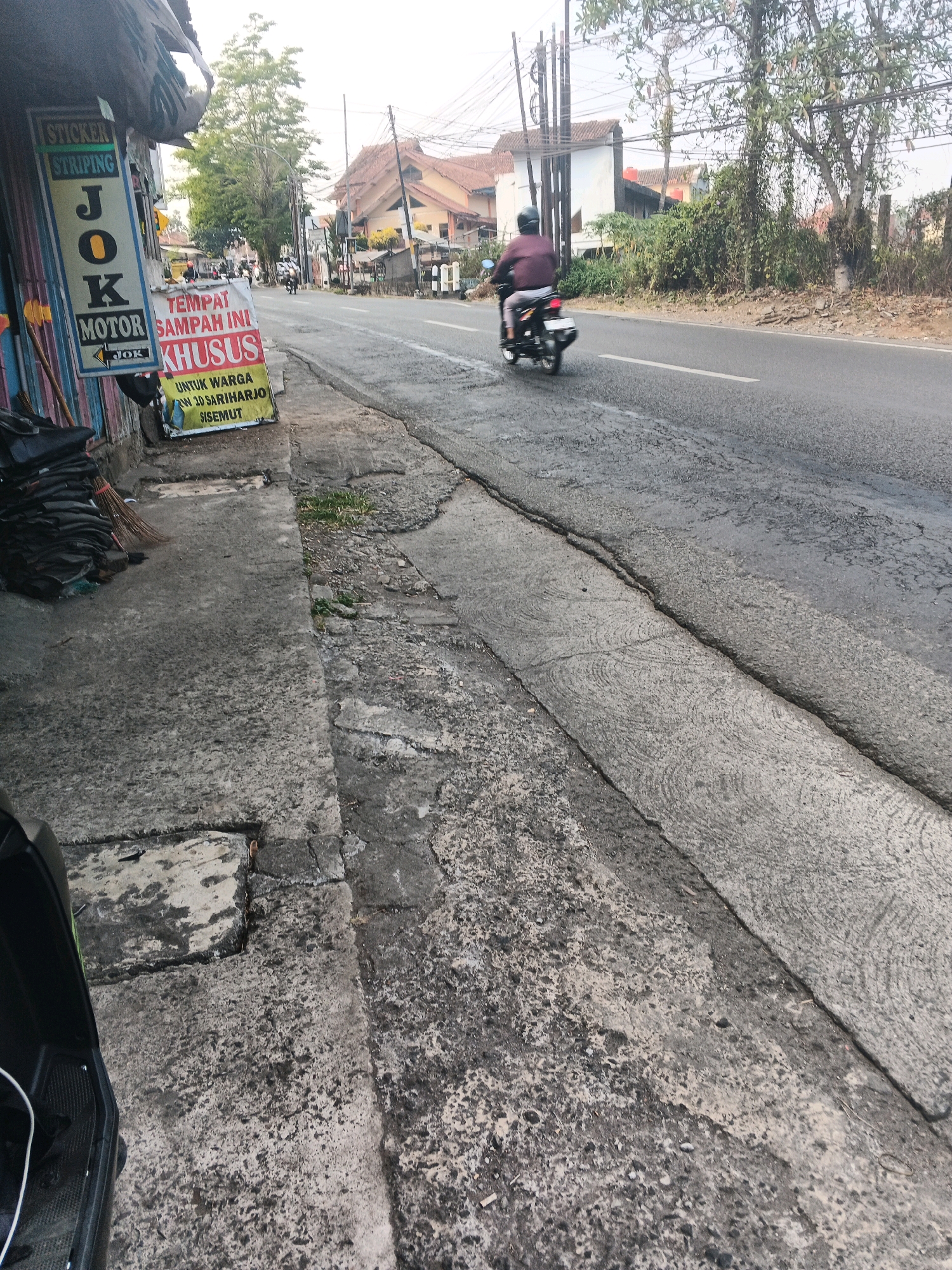 The image depicts a street scene in Ungaran, Indonesia. In the foreground, on the left, is a sidewalk with some stacked black items, possibly tires, and a broom. A sign is visible on a pole, with text that includes "STICKER STRIPING JOK MOTOR" and "TEMPAT SAMPAH INI KHUSUS UNTUK WARGA RW 30 SARIHARJO SISEMUT". This indicates a designated trash area for residents of the local RW (Rukun Warga) 30 in Sariharjo, Sisemut, Ungaran.
The street itself appears to be paved with asphalt and shows signs of wear and tear, with cracks and uneven surfaces. A motorcycle with a rider is traveling away from the camera down the street. In the background, there are buildings, trees, and utility poles with numerous wires, typical of a residential or mixed-use urban area. The sky is overcast, suggesting a daytime scene with diffused light. The overall impression is of a functional, everyday street in a moderately developed area.