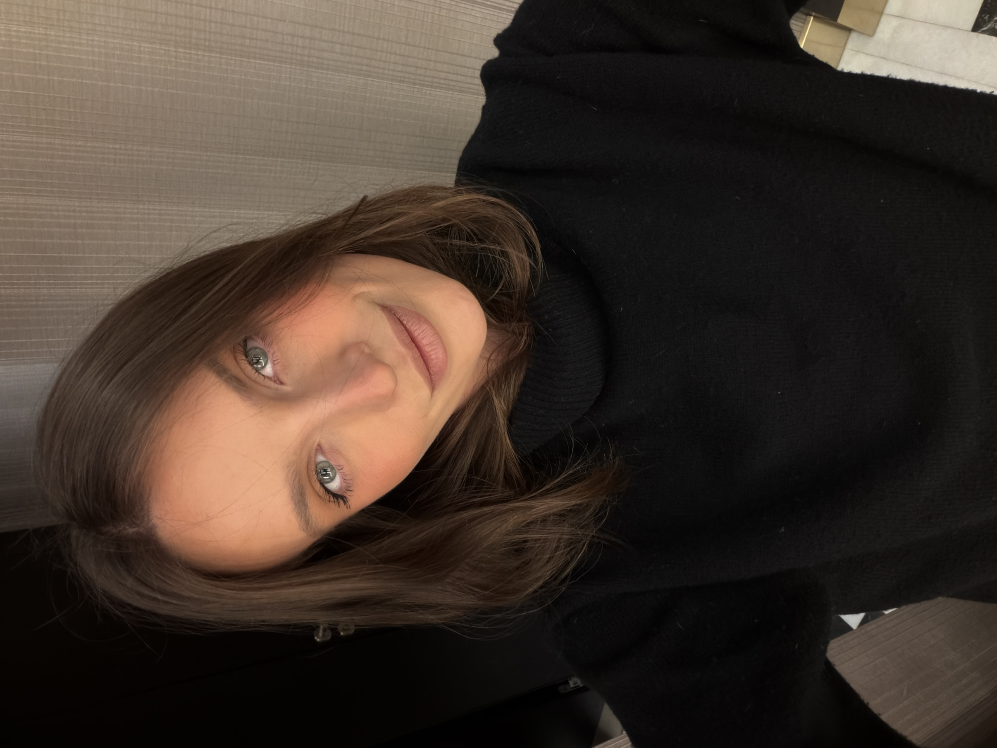 One female is visible in a close-up, head-and-shoulders indoor selfie taken in Budva, Montenegro. She has shoulder-length brown hair, light-colored eyes, and fair skin with subtle cheek blush and natural-toned lips. The subject wears a black turtleneck sweater and looks directly at the camera with a slight smile. The background consists of a dark, possibly paneled surface with a metallic handle on the left, and a vertically textured light-colored wall on the right. A tiled floor with a dark border is partially visible at the bottom right.