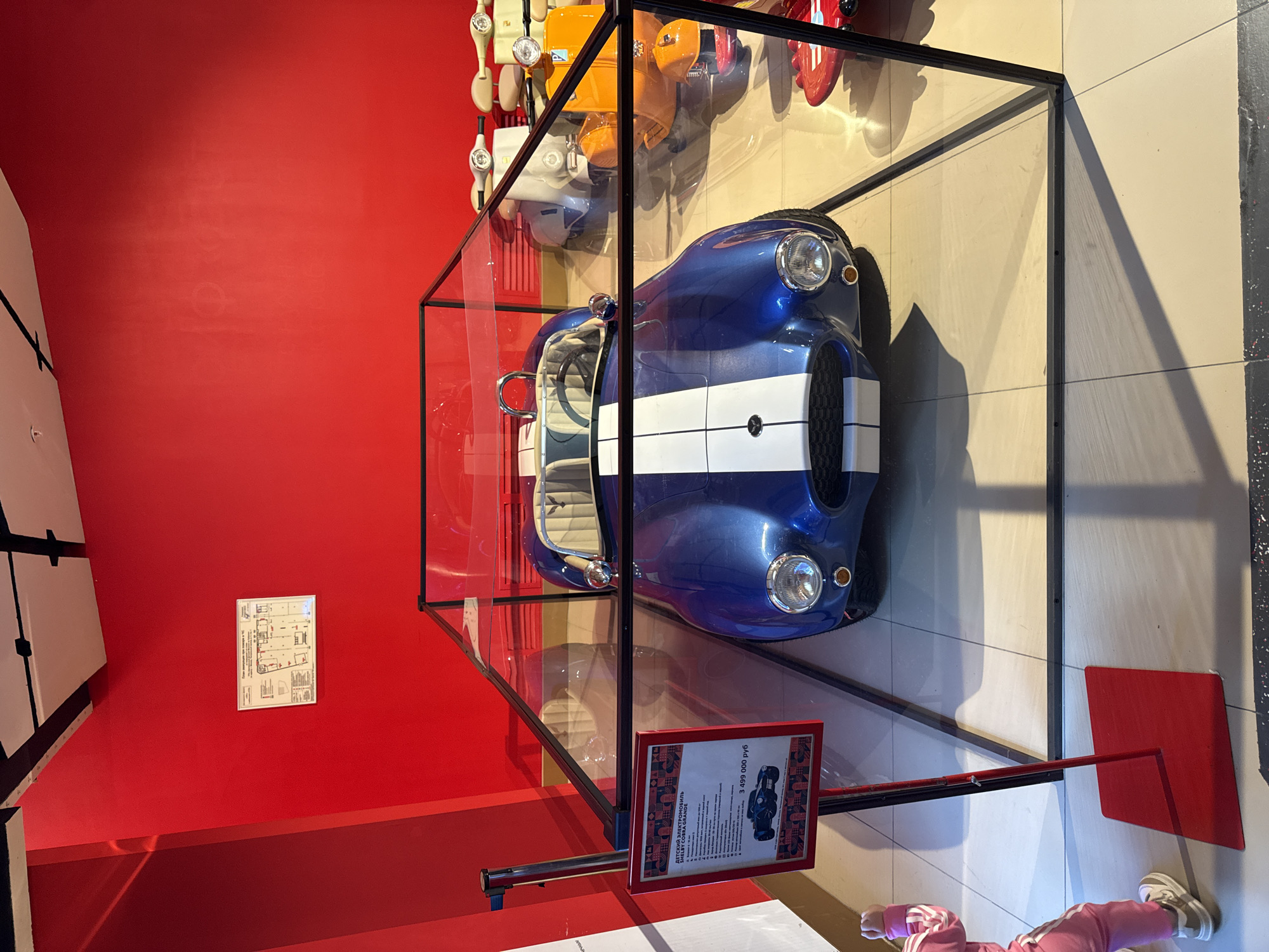 The image shows a blue car with a white stripe encased in a glass display case. The car appears to be a vintage model. The display is located in a room with a red wall. There is a sign in front of the case. The location is in Moscow, Russia. There is a person wearing pink pants and white shoes in the bottom right corner of the image.