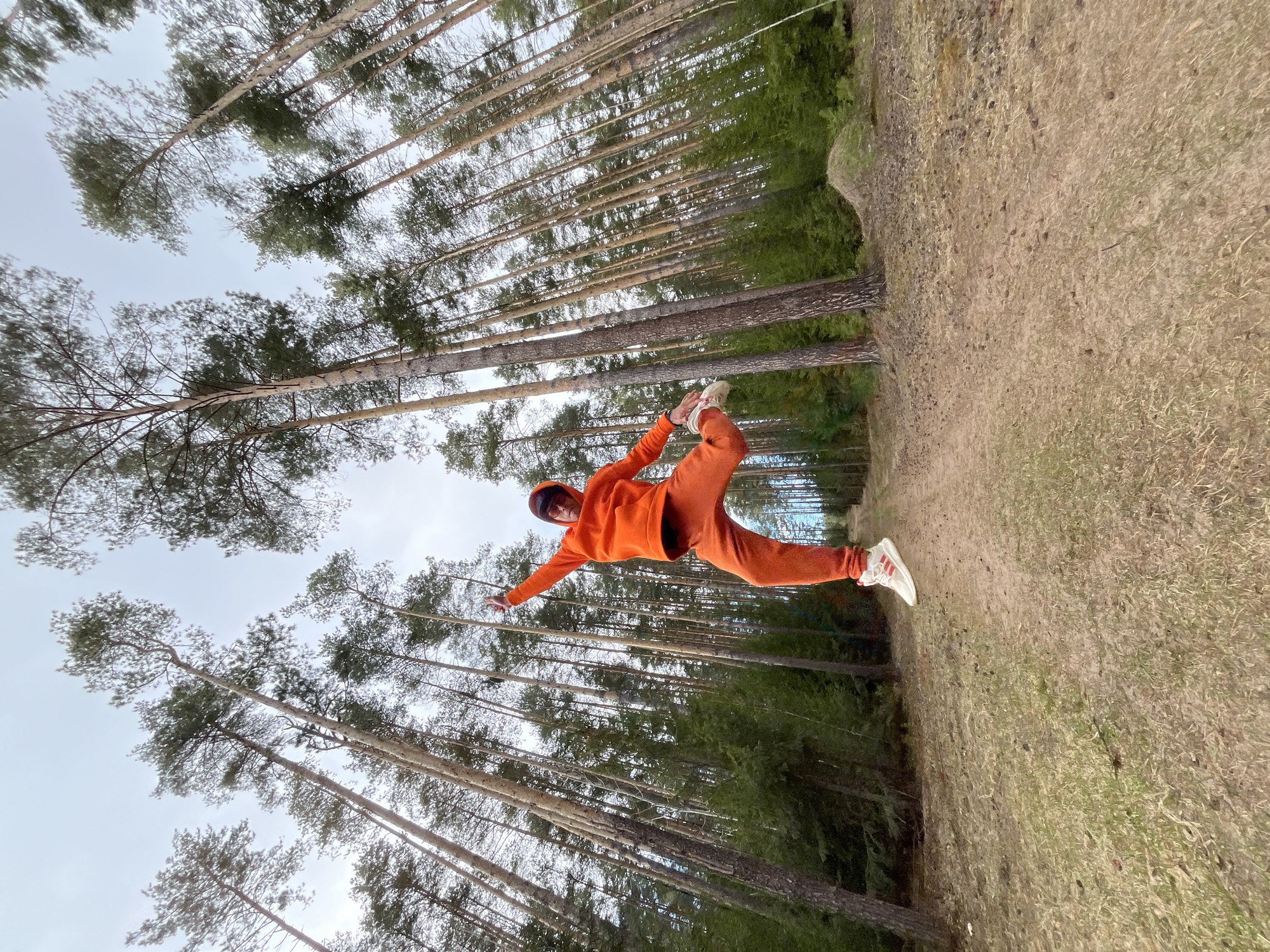An energetic, full-body outdoor shot captures a person in mid-air within a pine forest, likely in Chaikovsky, Russia. The individual, appearing to be male, is dressed in a vibrant, matching orange hooded tracksuit (hoodie and sweatpants) and white athletic shoes with three red stripes. They are depicted in a dynamic, acrobatic pose, with one leg bent at the knee and the foot positioned near the opposite leg's thigh, while the other leg extends backward and is slightly bent. Both arms are outstretched to the sides, conveying a sense of balance and motion.
The background is filled with numerous tall, slender pine trees with green foliage, suggesting a dense wooded environment. The ground beneath is covered with dry, light brown grass and some patches of green, indicating natural, unpaved terrain. The sky above is overcast, casting a soft, diffused light over the scene, suggesting a cloudy day during daytime hours. The overall impression is one of agility, freedom, and activity amidst natural surroundings. No explicit text is clearly visible on the clothing or shoes.
