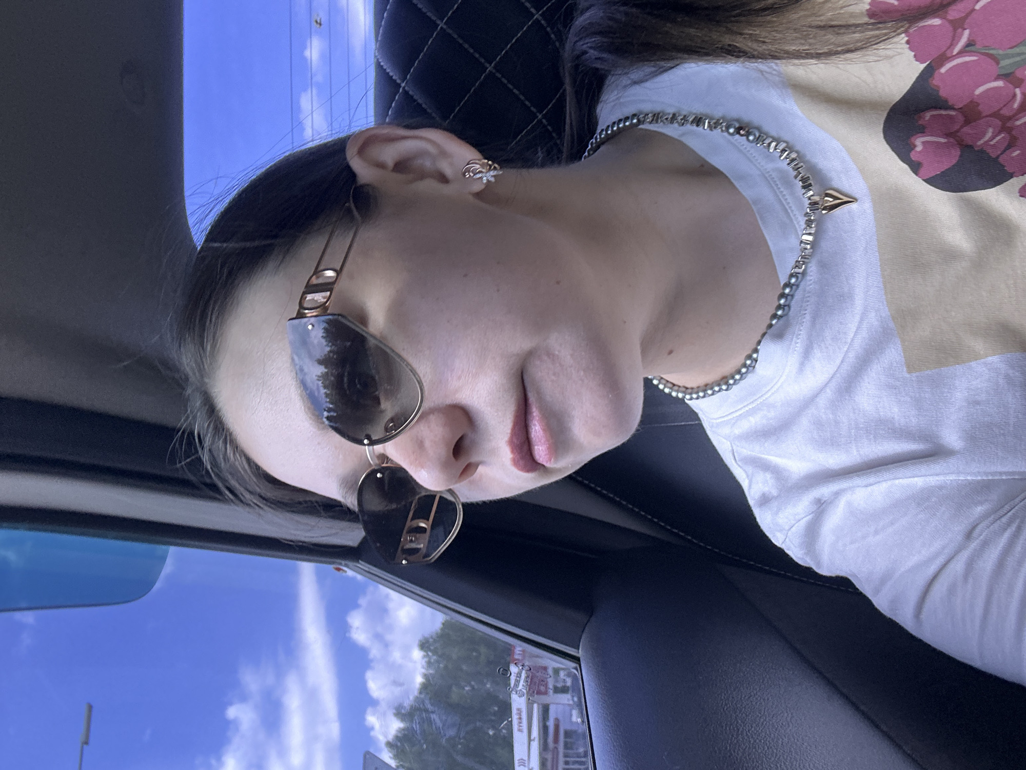 The photograph, taken inside a vehicle, shows a person wearing sunglasses and a necklace. The person has an earring in their left ear. The view through the car window reveals an exterior scene, with visible buildings and trees in the background. Based on the signs visible through the window, this appears to be Chaikovsky, Russia.