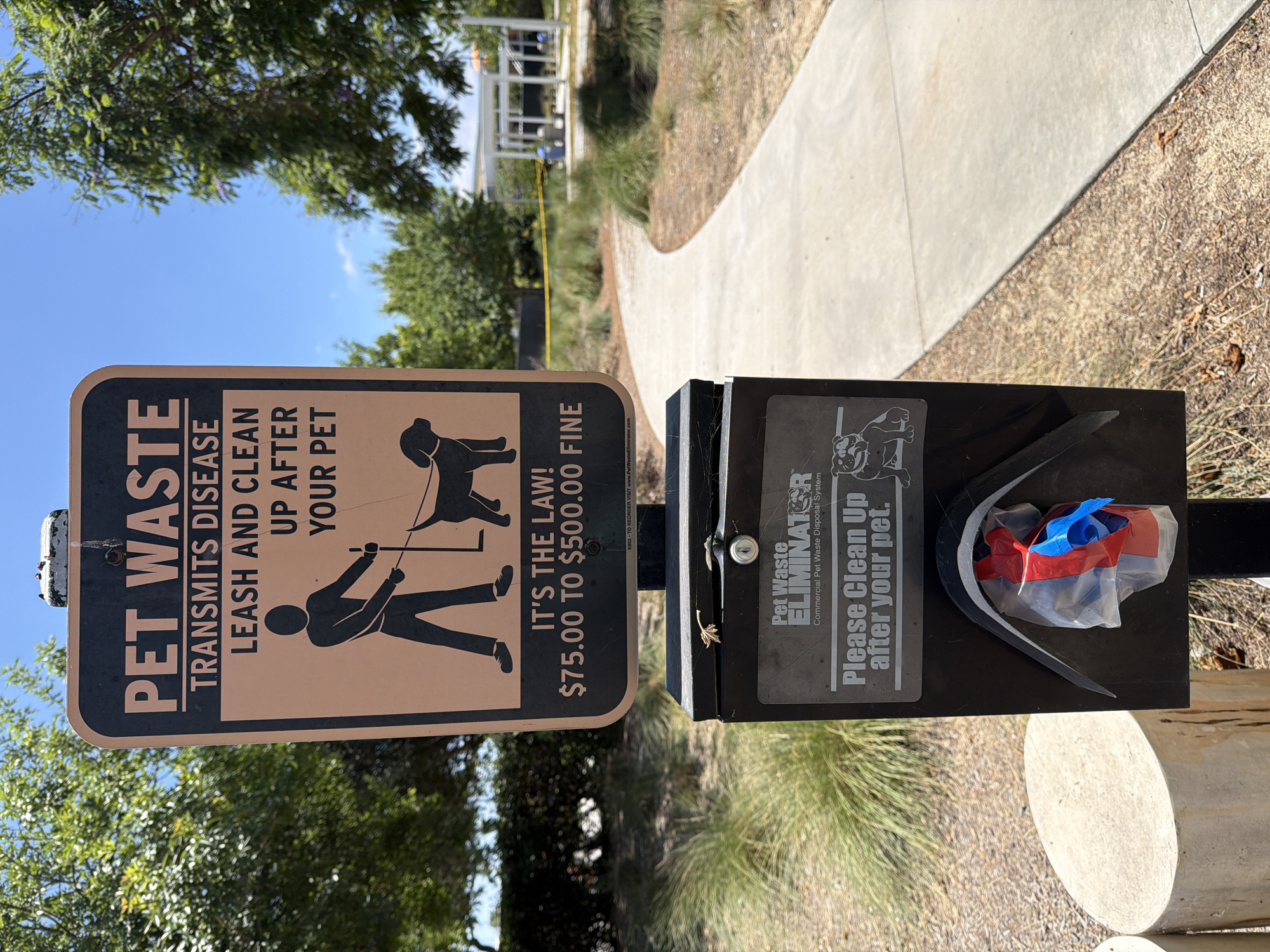 A pet waste station with a sign and dispenser is situated outdoors. The sign, featuring a silhouette of a person walking a dog and text that reads "PET WASTE TRANSMITS DISEASE LEASH AND CLEAN UP AFTER YOUR PET IT'S THE LAW! $75.00 TO $500.00 FINE," is mounted on a pole above the dispenser. The dispenser is a black metal box with a slot for waste bags and a chute where a plastic bag with red and blue ties is visible. To the left of the station is a light-colored, cylindrical object that appears to be made of concrete. A paved pathway curves to the right of the station, bordered by dry grass and some greenery. In the background, there are trees and a white structure with a yellow barrier in front of it, possibly indicating a park or public area. The sky is clear and blue. The location is the city of Irvine, United States of America.