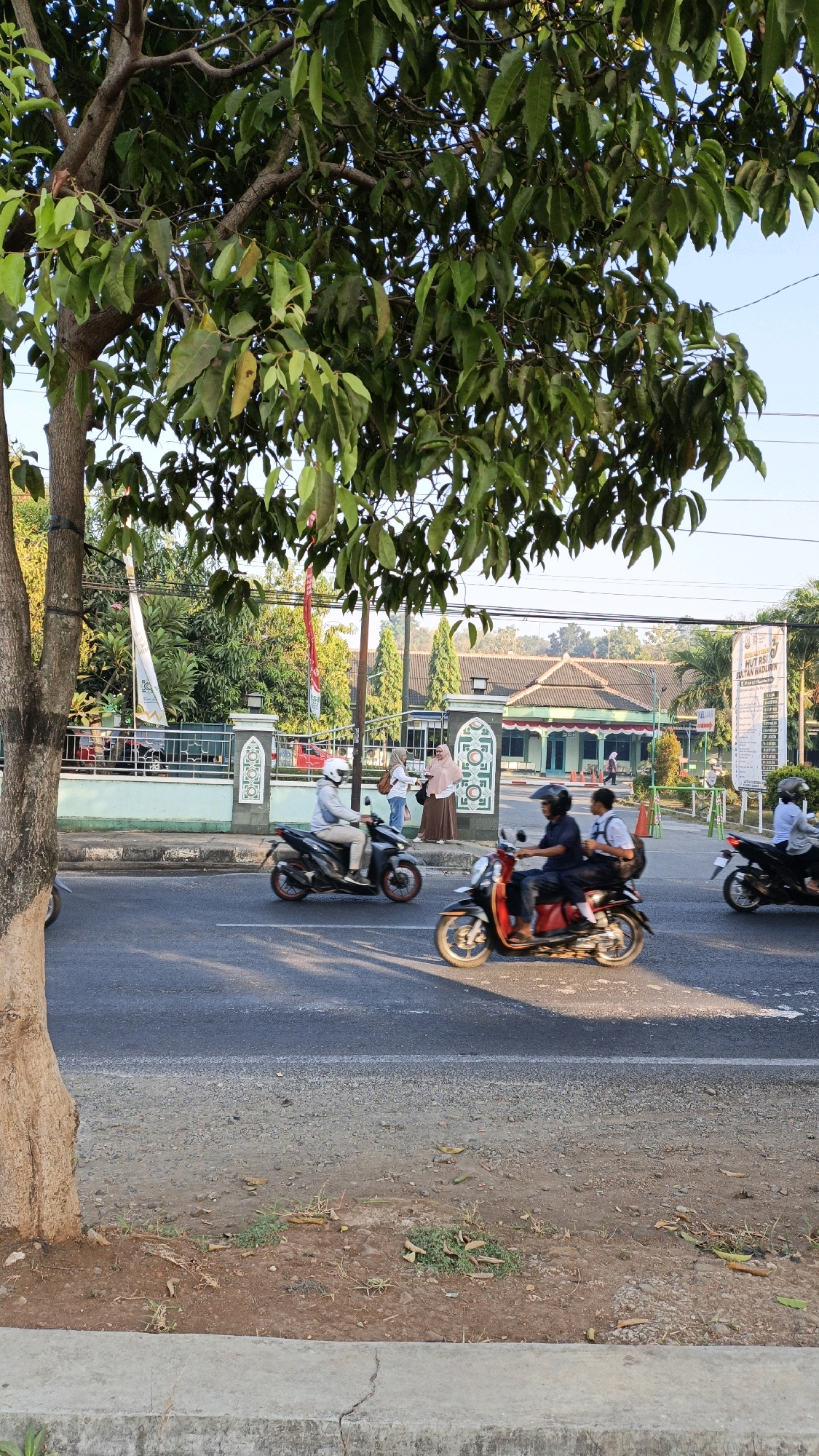 The image depicts a street scene in Kuwasen, Indonesia, during the daytime. In the foreground, a tree with abundant green leaves partially obscures the view of the road. The road itself is paved and appears to be a two-lane street with traffic moving in the same direction.
Several people are seen riding motorcycles. On the left, a person wearing a white helmet and a light-colored jacket is riding a dark-colored scooter. To their right and slightly further down the road, another scooter is visible with two individuals on it. The rider is wearing a dark shirt, and the passenger, a young person, is wearing a white shirt and dark pants, possibly a student. Further to the right, a third motorcycle is partially visible with its rider wearing a helmet.
On the sidewalk bordering the road, there are pedestrians. Two women are standing and interacting near the entrance of a building complex. One is wearing a light-colored hijab and a brown dress, while the other wears a white hijab and a light blue top. Behind them, other pedestrians can be seen walking.
The background reveals a building with a green roof and an arched entranceway adorned with decorative patterns. Flags are also visible, including Indonesian flags and other banners. Power lines are present in the upper portion of the image. The overall environment suggests a typical urban or suburban street in Indonesia, with elements of local architecture and daily life. The lighting indicates it is likely late afternoon, given the shadows.