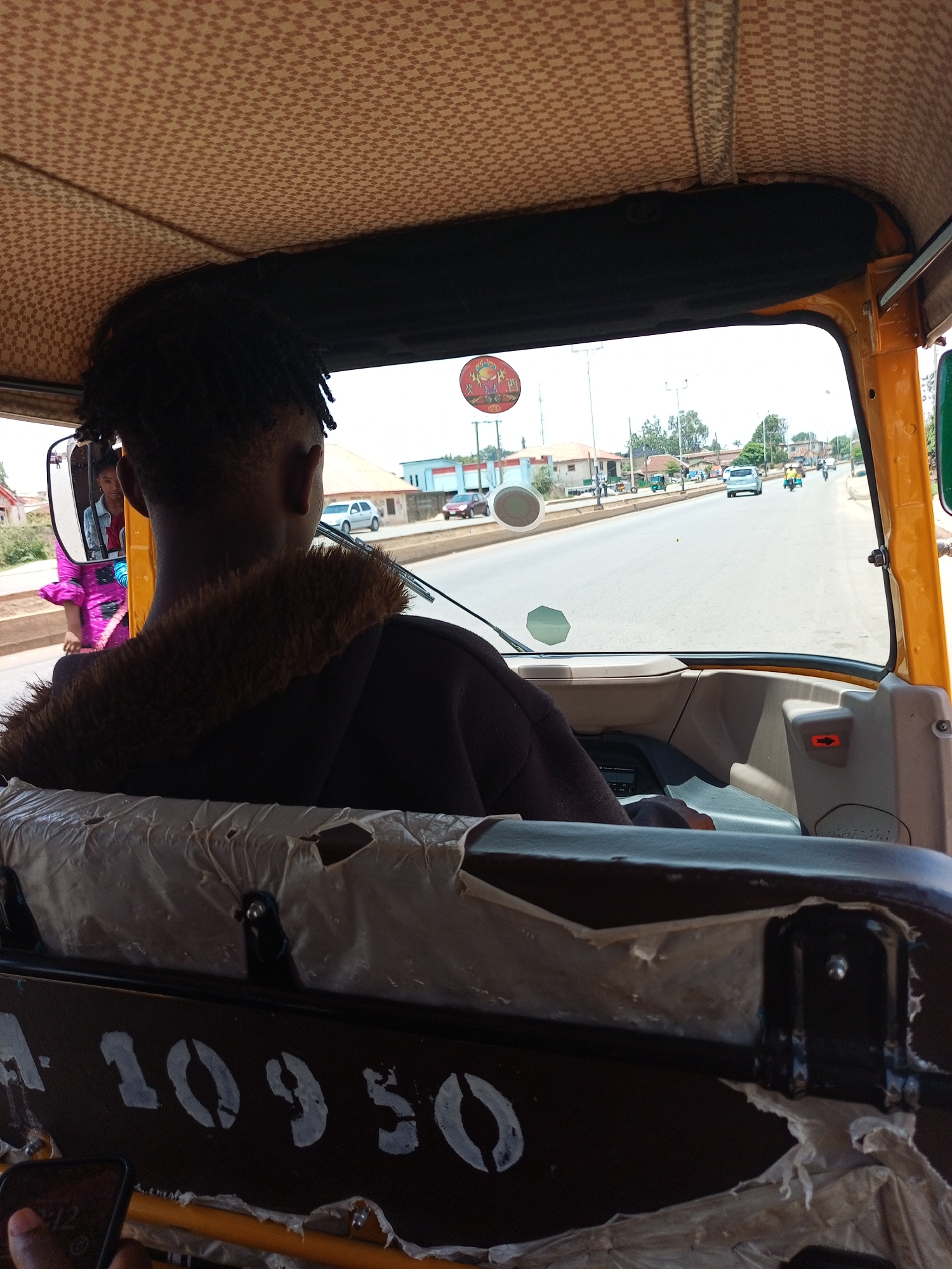The image, taken from inside a yellow tuk-tuk or auto-rickshaw, shows the driver with dark hair, facing forward. The road ahead shows other vehicles and buildings in Bukuru, Nigeria. A passenger is visible in the side mirror. The seat in front of the passenger is damaged. The writing on the back of the seat reads "-10950".
