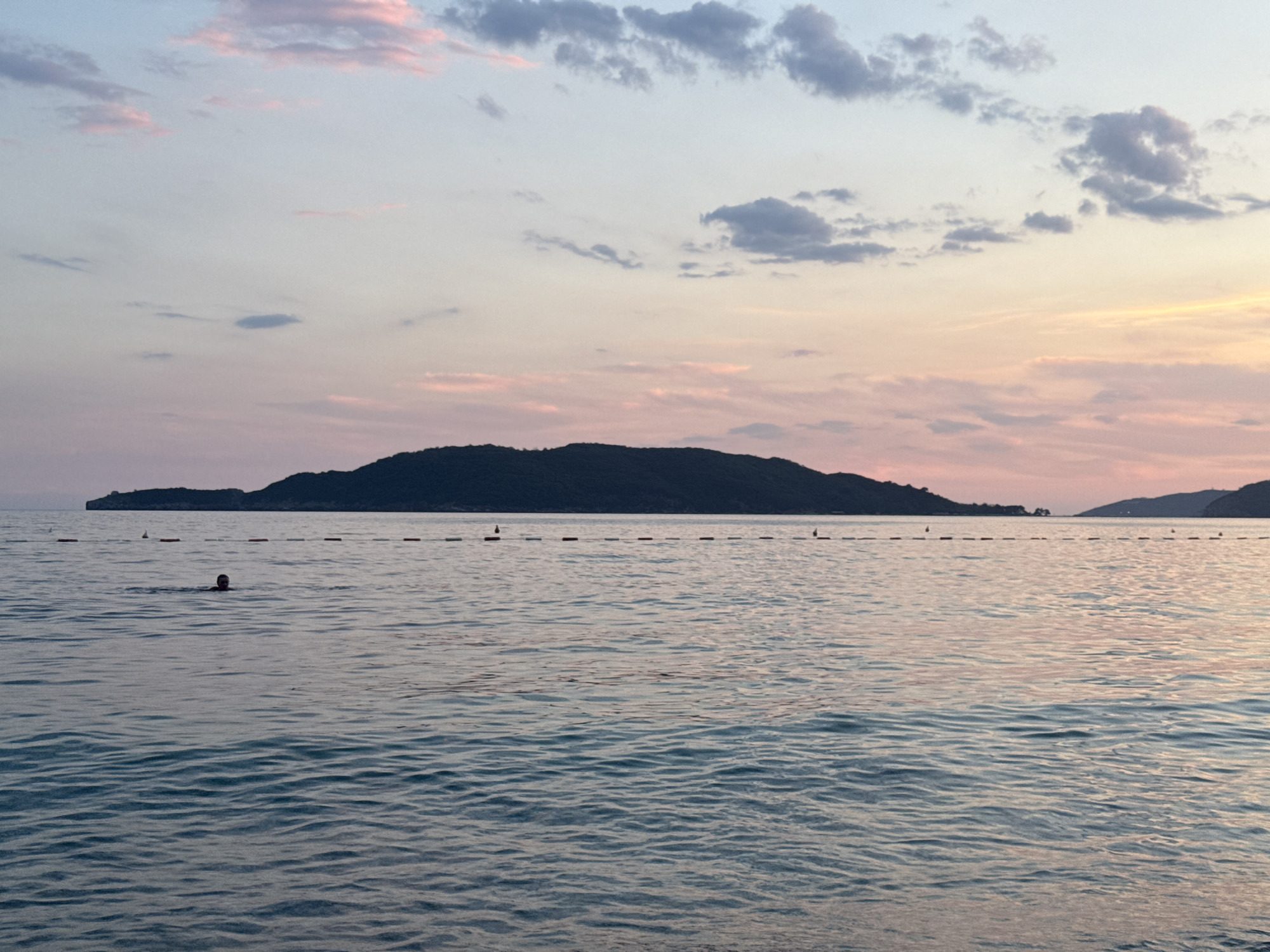 The image shows a serene seascape at dusk, likely from the beach in Kamenovo, Montenegro. The sky is painted with soft hues of pink, orange, and pale blue, with scattered clouds reflecting the fading sunlight. In the foreground, calm turquoise water gently laps against the shore, with subtle ripples creating a textured surface. A boundary line marked by buoys stretches across the water, indicating a designated swimming area. In the middle ground, a lone swimmer is visible, their head and shoulders breaking the surface of the water as they swim peacefully. In the background, a dark, silhouetted landmass, possibly an island or the mainland coastline, rises from the horizon, adding depth and a sense of natural grandeur to the scene. The overall atmosphere is one of tranquility and natural beauty.