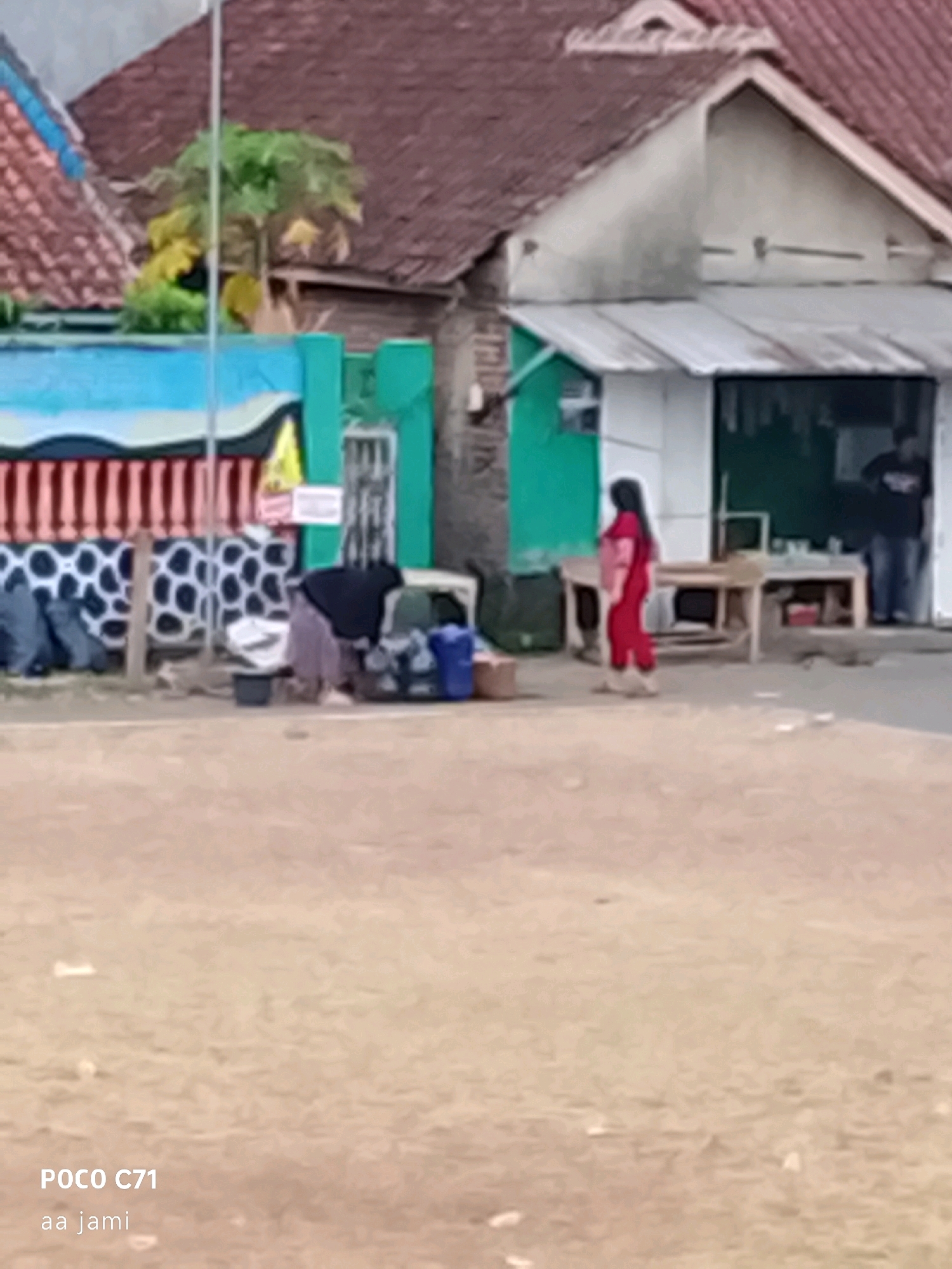 The image depicts an outdoor scene in what appears to be a residential or commercial area in Tasikmalaya, Indonesia, as indicated by the text overlay "Tasikmalaya". In the foreground, a sandy or dirt ground is visible with some debris scattered on it. In the mid-ground, a person dressed in a dark headscarf and a greyish-purple garment is bent over, appearing to be sorting or handling items. Next to this person are various objects, including what look like bags, containers, and possibly laundry items. A blue plastic container is also present. To the right of this person, another individual wearing a red outfit is standing and looking towards the left. Behind these individuals, a building with a green facade and a white-tiled roof is visible, with a portion of a roof made of reddish tiles in the background. A wooden table or counter is positioned in front of the green building. Further to the right, another person in dark clothing is standing near an open doorway. The overall atmosphere suggests a casual, possibly domestic or small business setting.