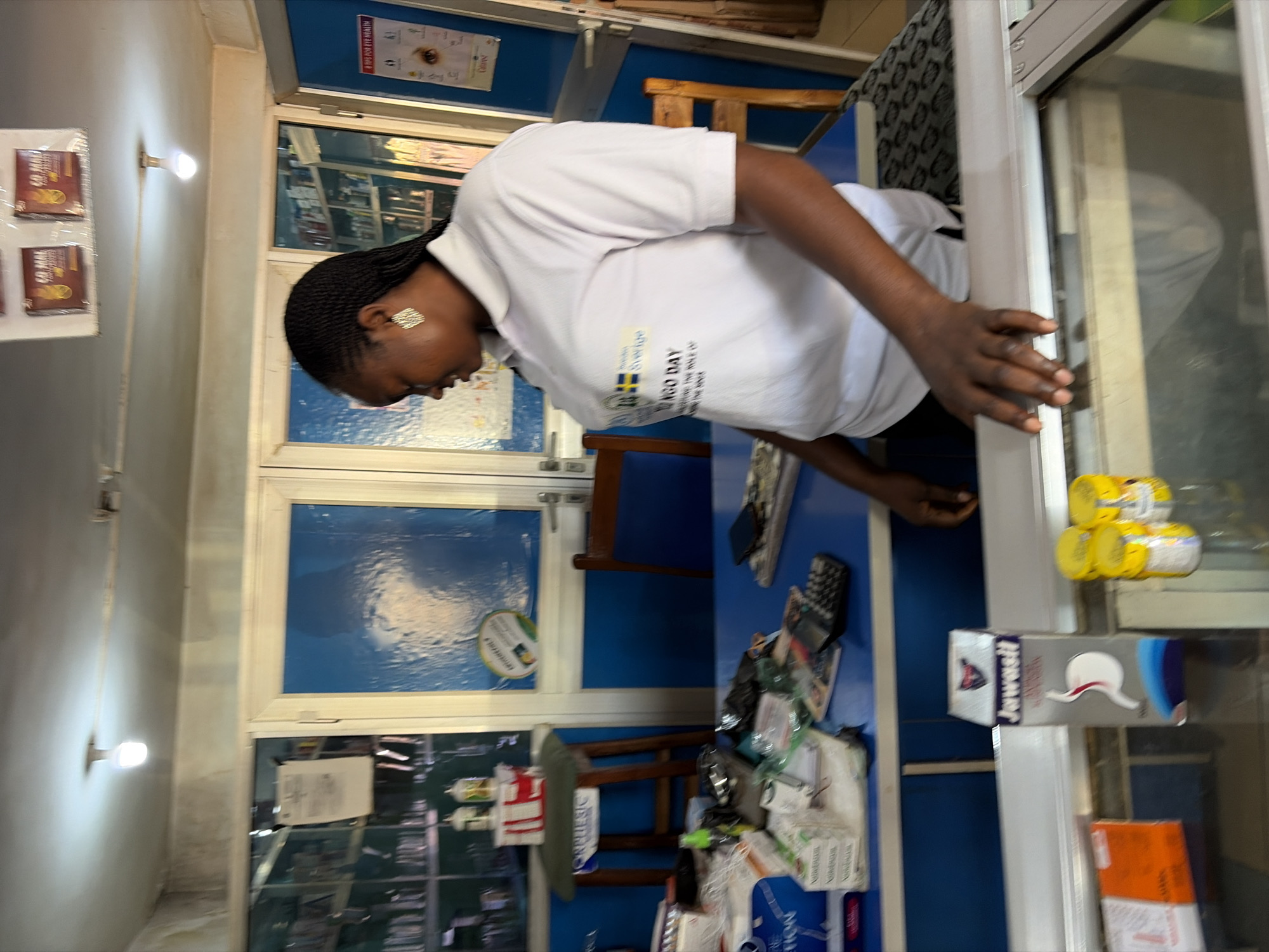 In a pharmacy in Jos, Nigeria, a Black woman with braided hair is standing behind a counter. She is wearing a white polo shirt with a logo on it, which appears to say "SWEDEN Sverige" and "NGO DAY". On the counter in front of her, there are several items, including boxes of medication, a calculator, a mobile phone, and a bottle of what looks like hand sanitizer. Behind her, shelves are stocked with various pharmaceutical products, and the room has blue walls and white doors. The overall scene suggests a busy and functional pharmacy environment.