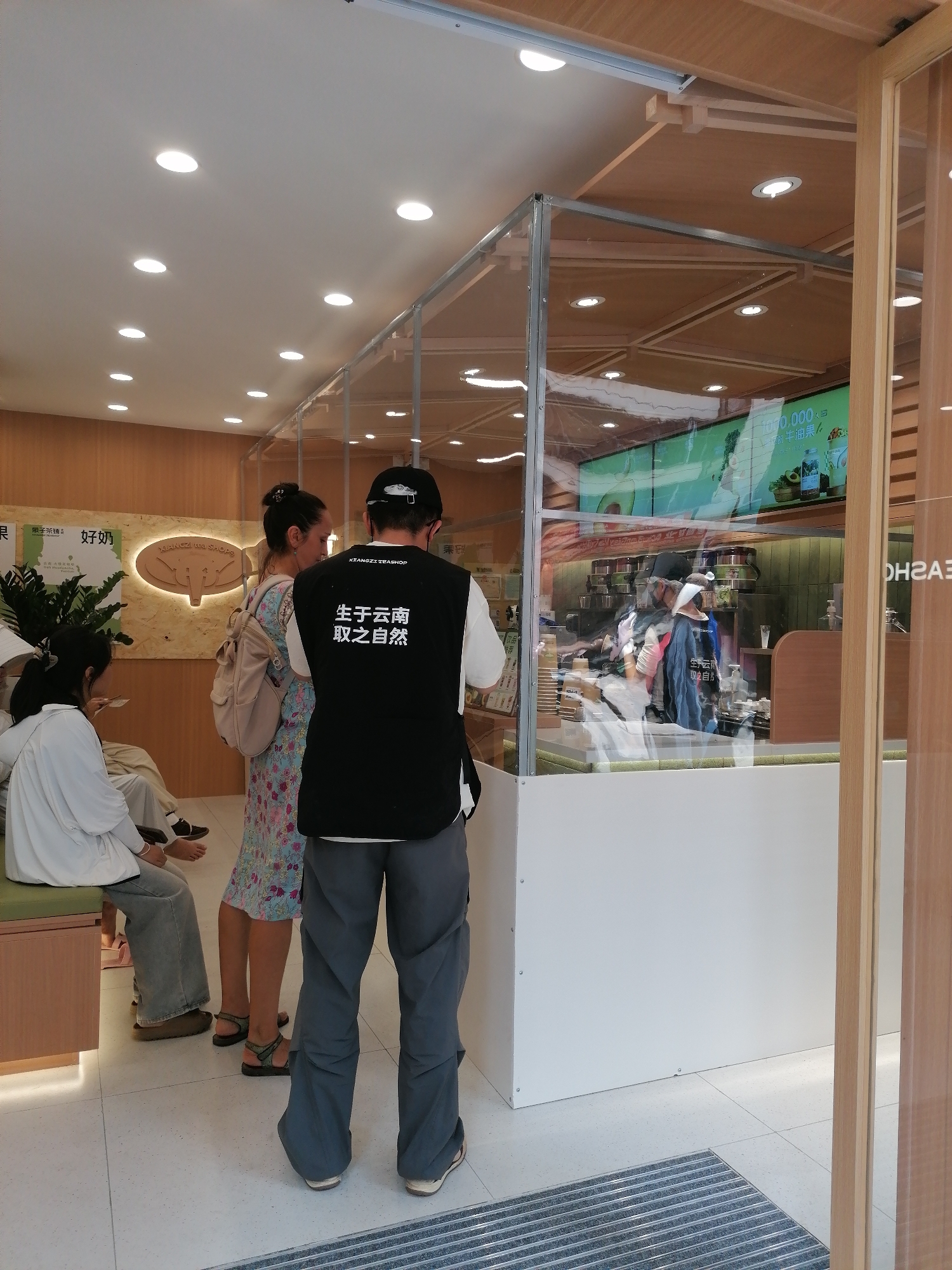 Here is a matter-of-fact description of the image:
The image shows an interior scene, possibly a shop or café, located in Hekou, China. Patrons are visible, one standing and facing the cashier area with his back towards the viewer, wearing a black vest with white Chinese characters on the back, grey pants, and a baseball cap. Another woman is standing beside him, looking at the cashier area and wearing a floral dress and carrying a backpack. Other people are seated on a low bench, near a potted plant.
The cashier area is encased in a clear, glass-like partition. The background wall is made of natural-colored wood. There's a lit sign with Chinese characters and an illustration of an elephant head on the wall. Signage is visible, featuring text related to tea and natural ingredients. The scene is brightly lit with overhead lighting fixtures. A doormat sits in front of what appears to be a doorway.
