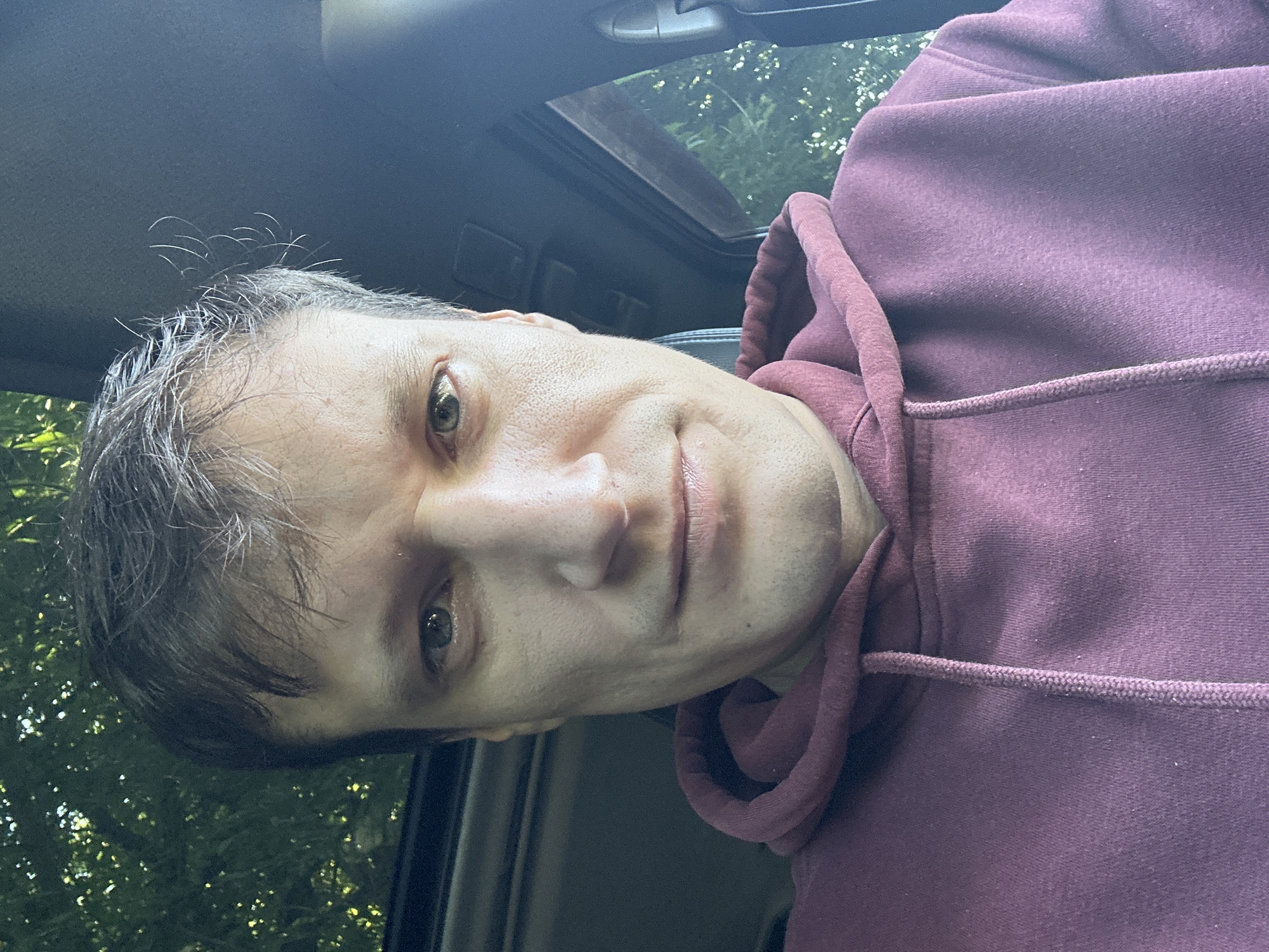 A close-up selfie of a man wearing a maroon hoodie. The man is looking directly at the camera with a neutral expression. He has short, dark hair with some gray strands. The background shows trees and what appears to be the interior of a vehicle, possibly with a sunroof. The image does not provide any information about the city of Makiv, Ukraine.