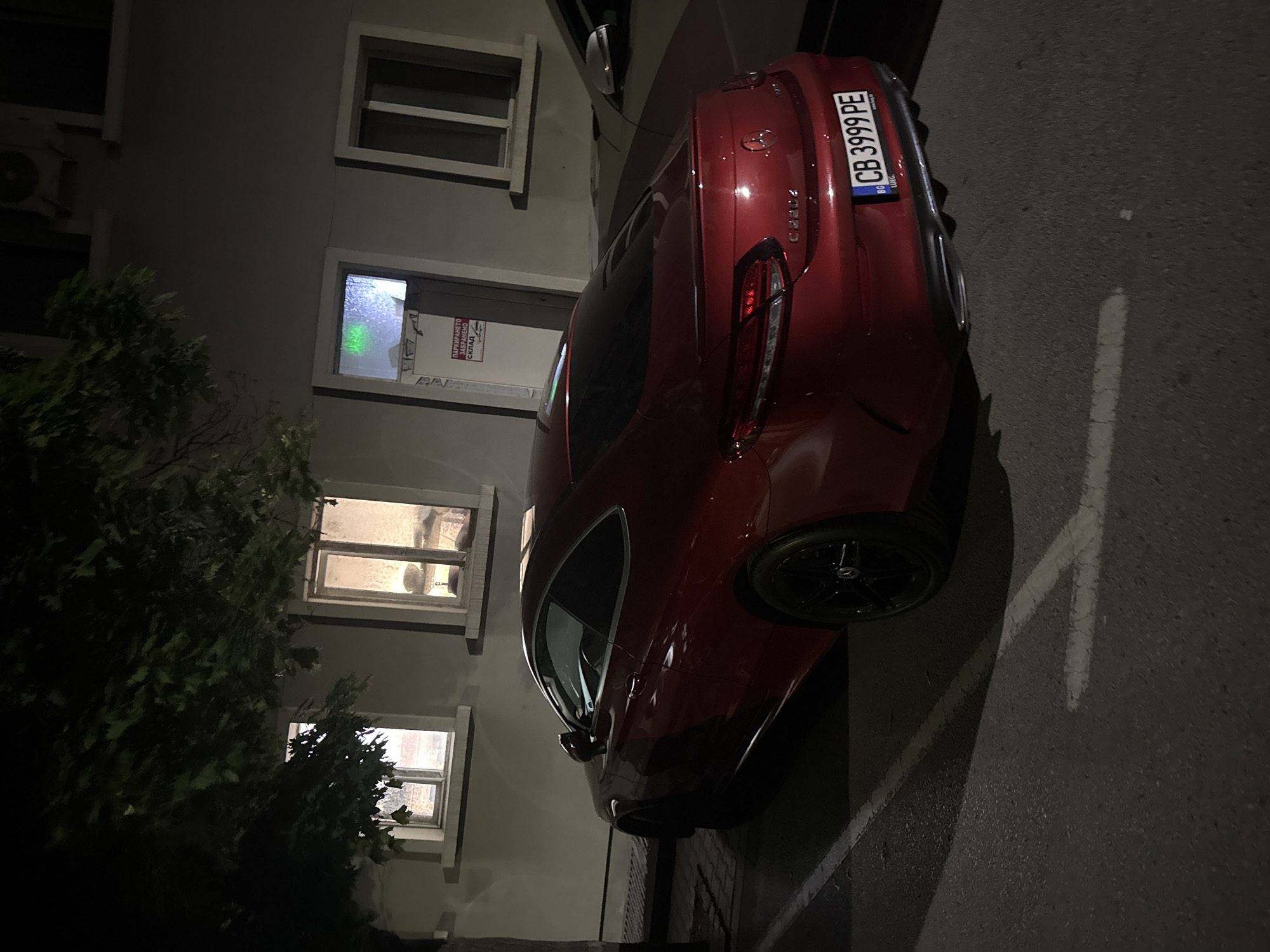 The image shows a red car with license plate "CB 3999 PE", parked on an asphalt surface in front of a building. The building is light-colored with multiple windows. A bush is visible on the left side of the frame. The location could be Blagoevgrad, Bulgaria, based on the license plate prefix "CB". The scene appears to be at night, with limited lighting. There are no people visible in the image.