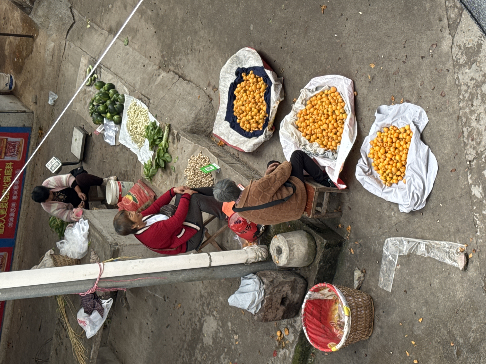 This aerial view depicts a street market scene in Hechuan, China. The market is set up on a concrete surface, possibly a sidewalk or public area. Several vendors are present, indicated by the assortment of produce and individuals seated nearby.
Three large bags, white with yellow fruit, are prominently displayed. To the left, other items include green gourds, long beans, and small piles of nuts. Two women are visible, one in a red sweater and another in a brown jacket, sitting on stools near their wares. Another woman is crouched on the ground near the green produce.
Additional objects include a weighing scale, several baskets, and containers. Litter, including plastic bags and scattered fruit remnants, is visible on the ground, suggesting a busy trading environment. A pipe runs horizontally across the scene, and a sign is partly visible on the left.