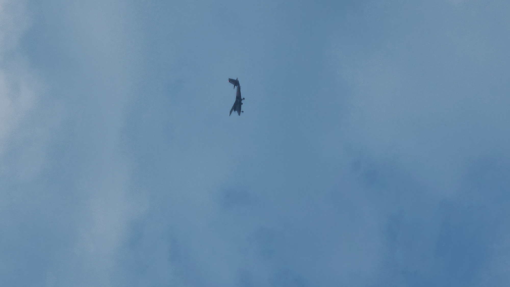An airplane is flying in a hazy blue sky. The airplane is a twin-engine prop plane, viewed from below and slightly to the side. It has a dark fuselage with a red stripe on its tail. The plane is positioned in the upper right quadrant of the image. The sky is overcast with faint cloud formations, indicating it is likely daytime. The scene does not contain any identifiable landmarks or features that would specifically place it in Castelló d'Empúries, Spain.