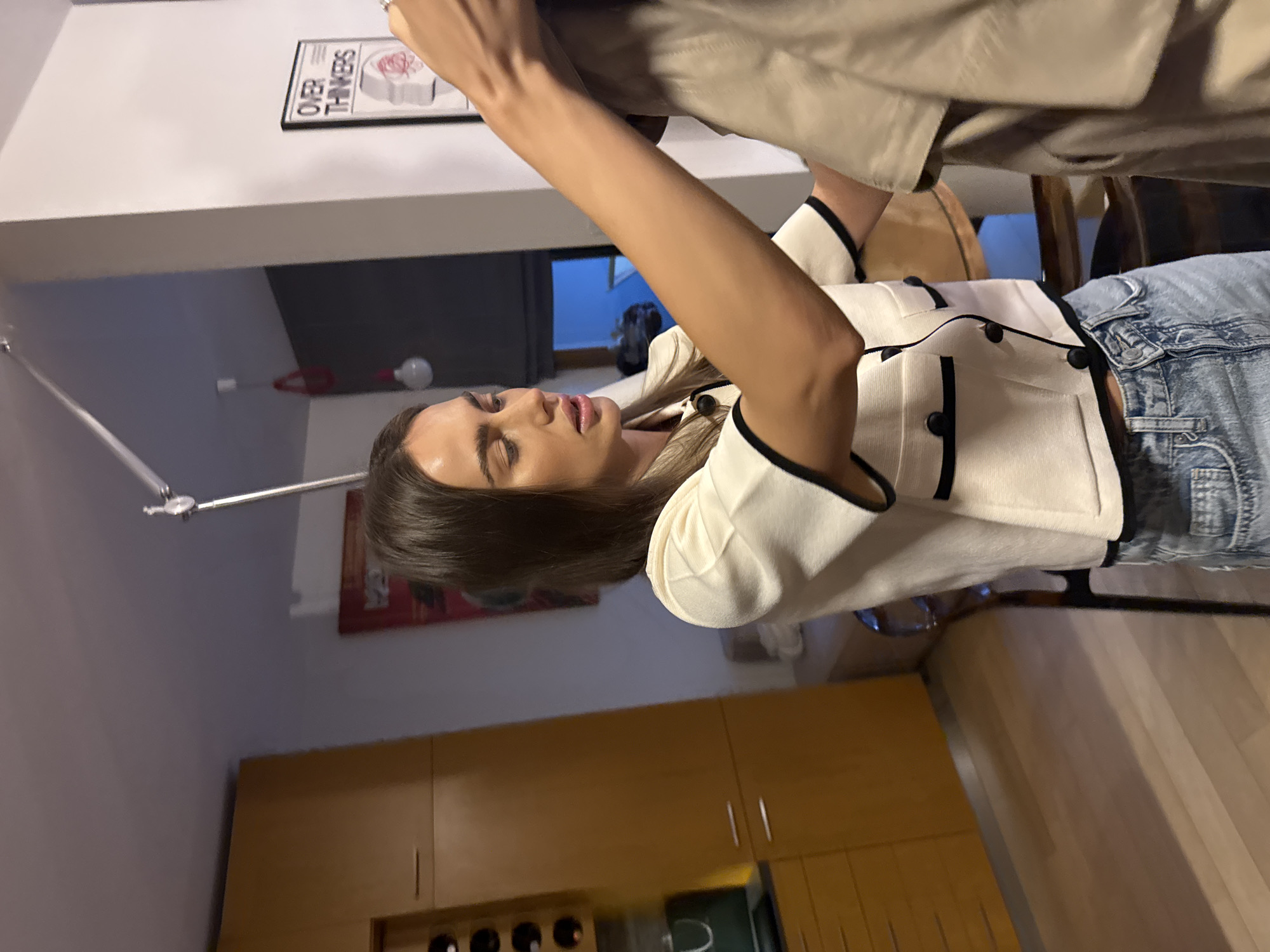A woman is captured indoors, wearing a cream-colored top with black trim and blue denim jeans. She is partially obscured by a person holding up a khaki-colored jacket or shirt. The background shows kitchen cabinets with various objects on the countertop, a white ceiling, and a wall with a poster that reads "OVER THINKERS." In the distance is a window with what appears to be another building visible. The location seems to be a residential kitchen.